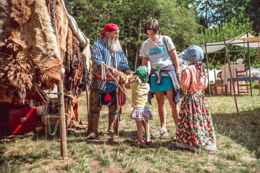 A bearded man in period costume and red cap interacts with a woman and child at an outdoor encampment with furs and tents.