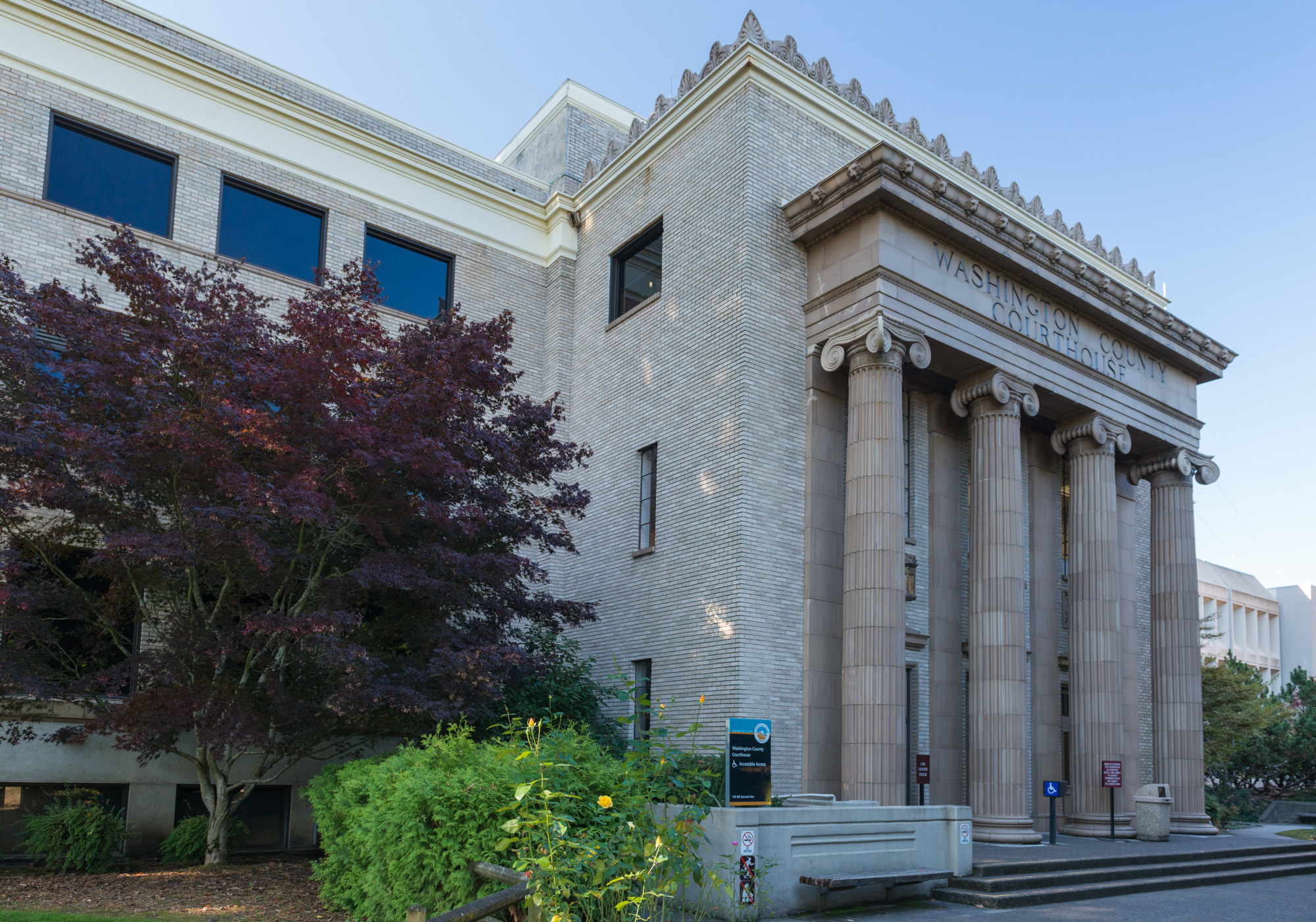 Stone courthouse portico with tall Ionic columns and a red-leafed tree in front.