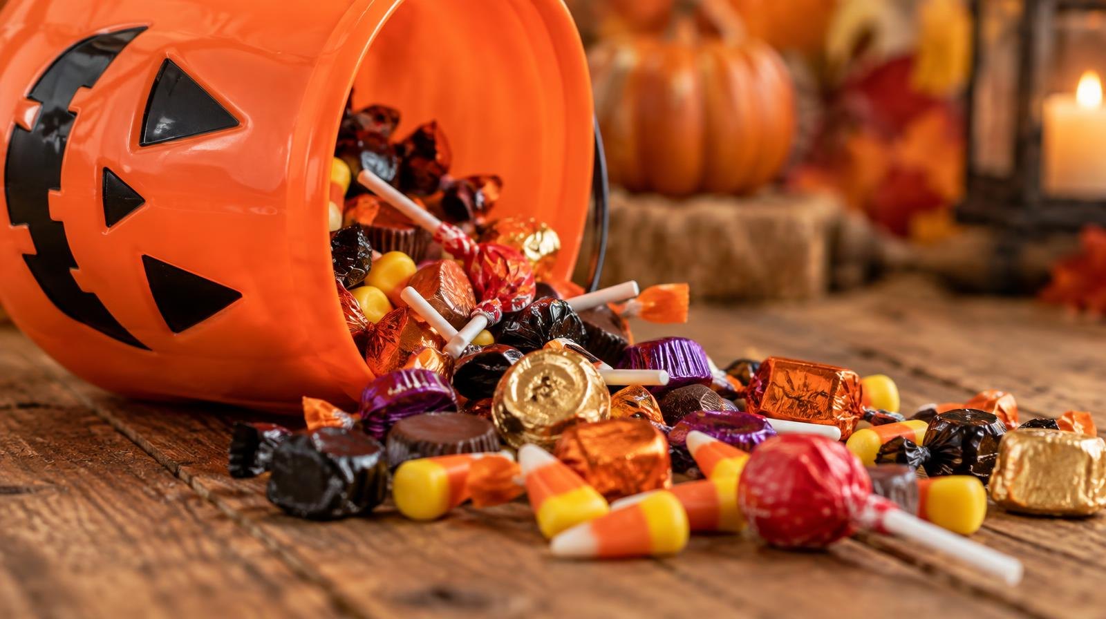 Orange pumpkin bucket overflowing with colorful Halloween candy on a wooden surface.