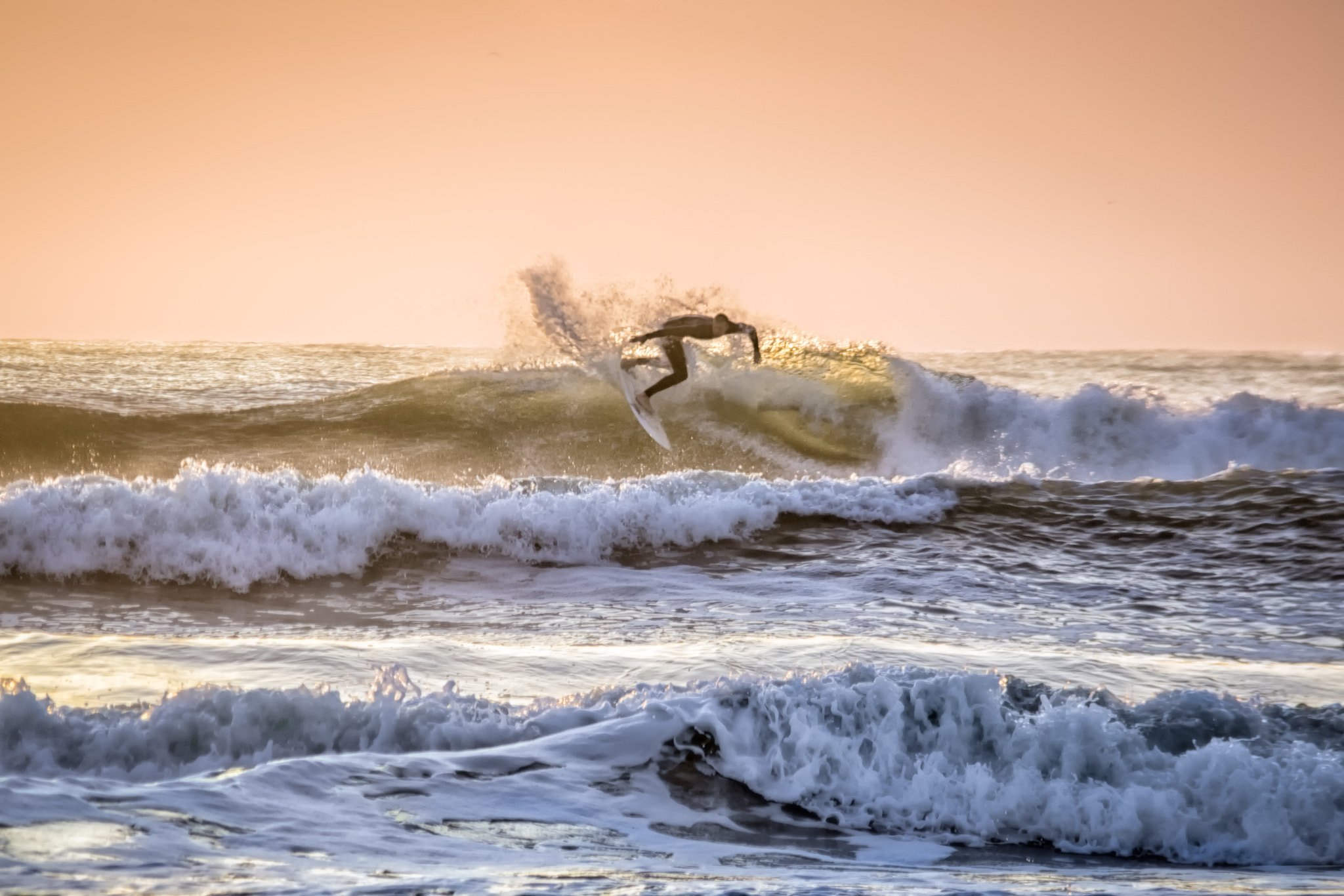 Surfer riding a wave near a pier at sunrise with orange sky.