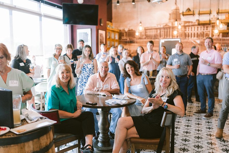 Crowd of adults socializing inside a warmly lit pub with Edison-bulb lighting.