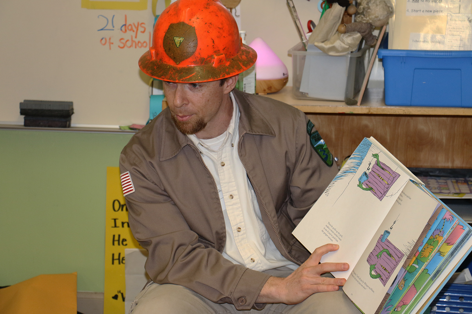 A man in a hard hat reads a picture book to young children in an elementary classroom.