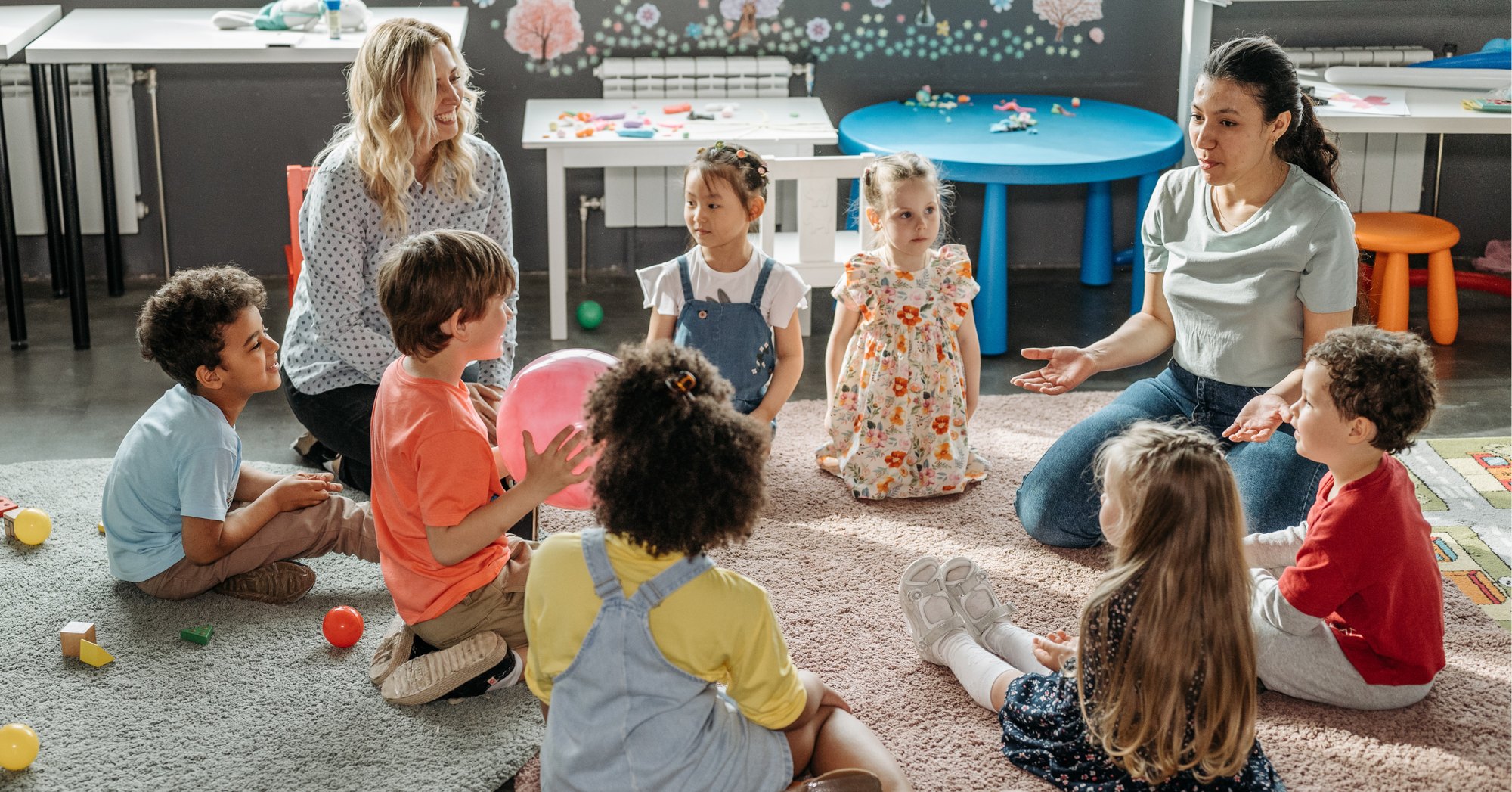 Two women sit with young children in a circle on a rug in a colorful classroom.