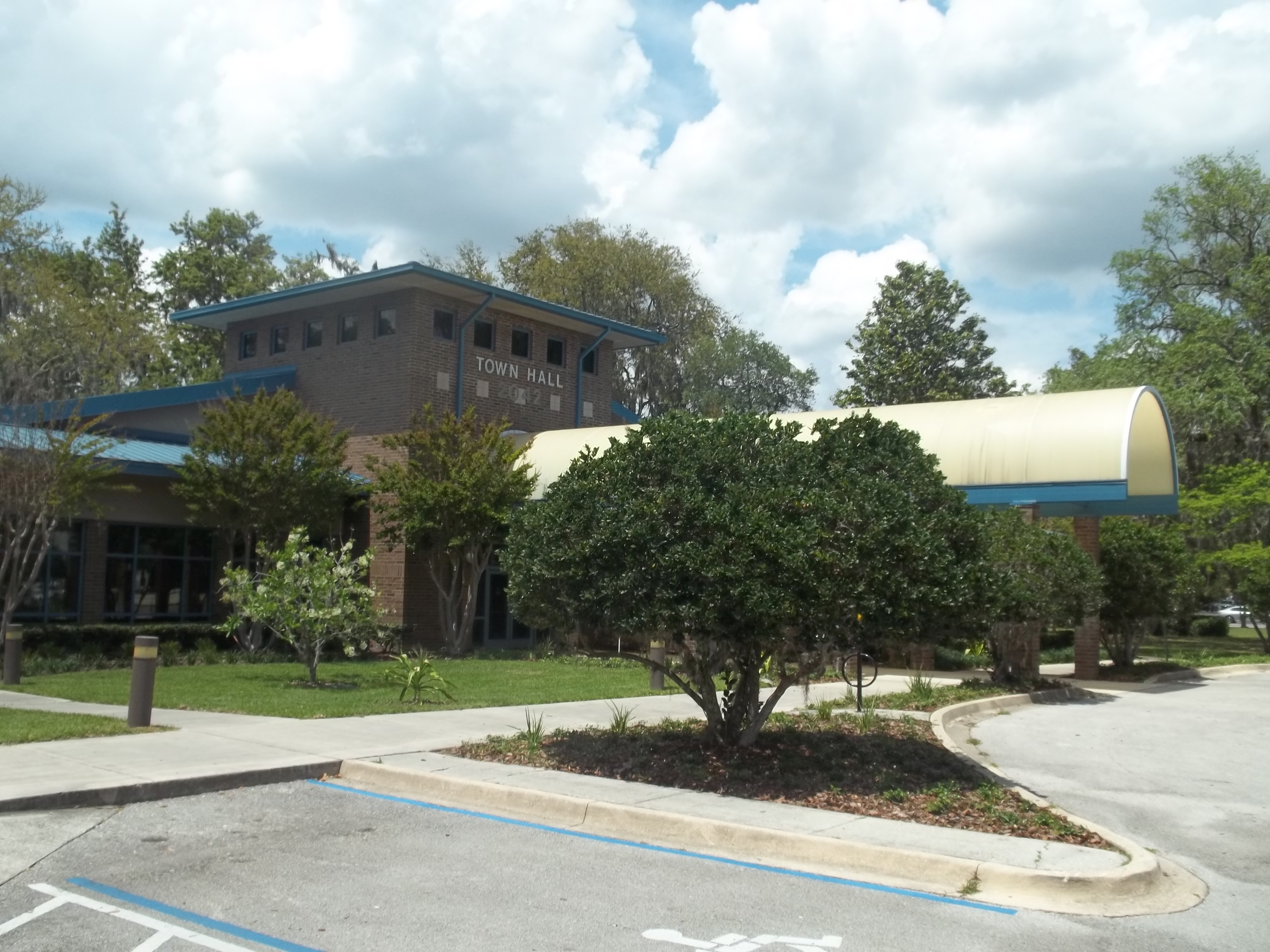 Brick municipal building with blue trim and yellow canopy entrance, surrounded by trees.
