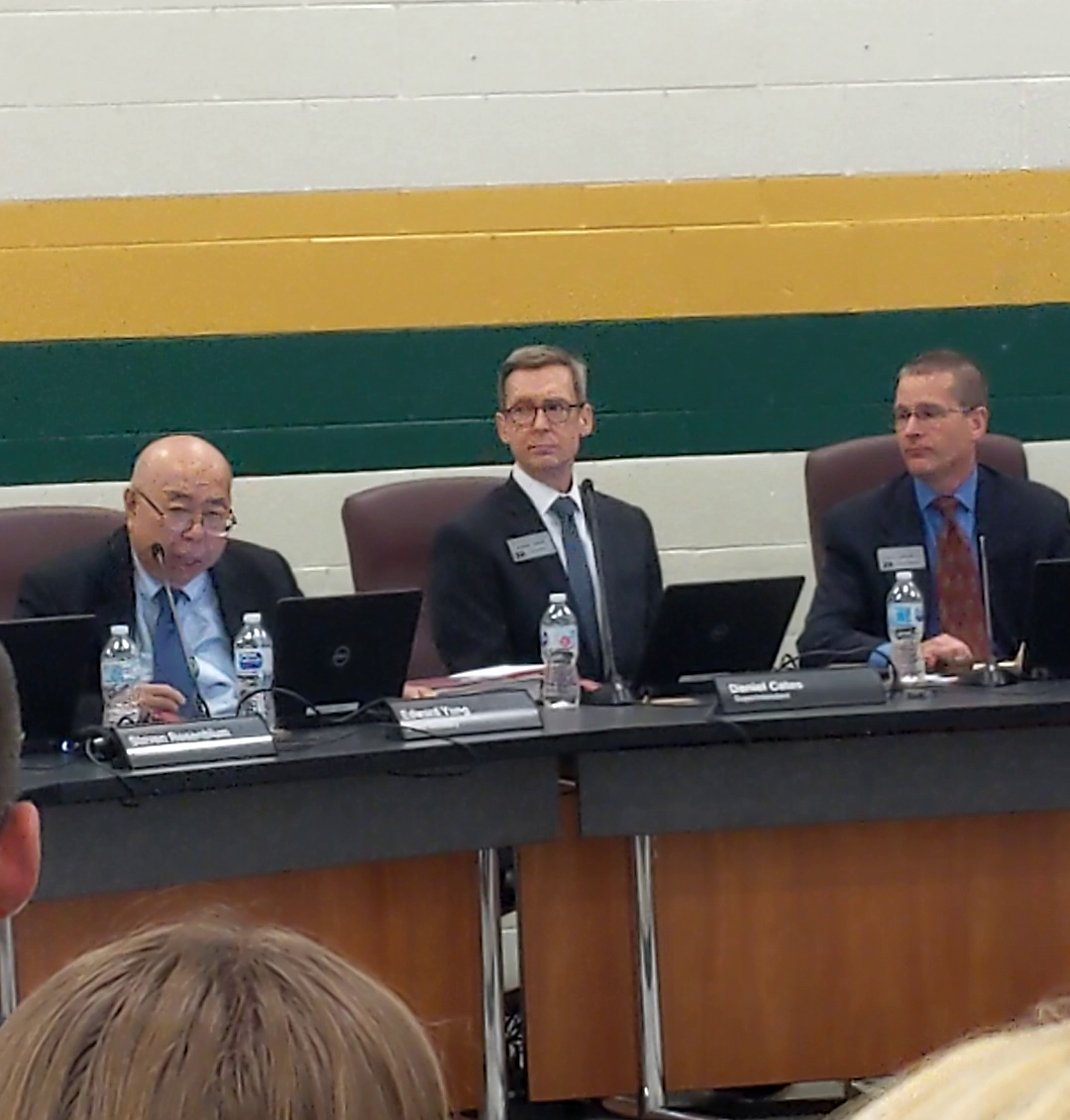 Three officials in suits seated at a long table during a school board meeting.