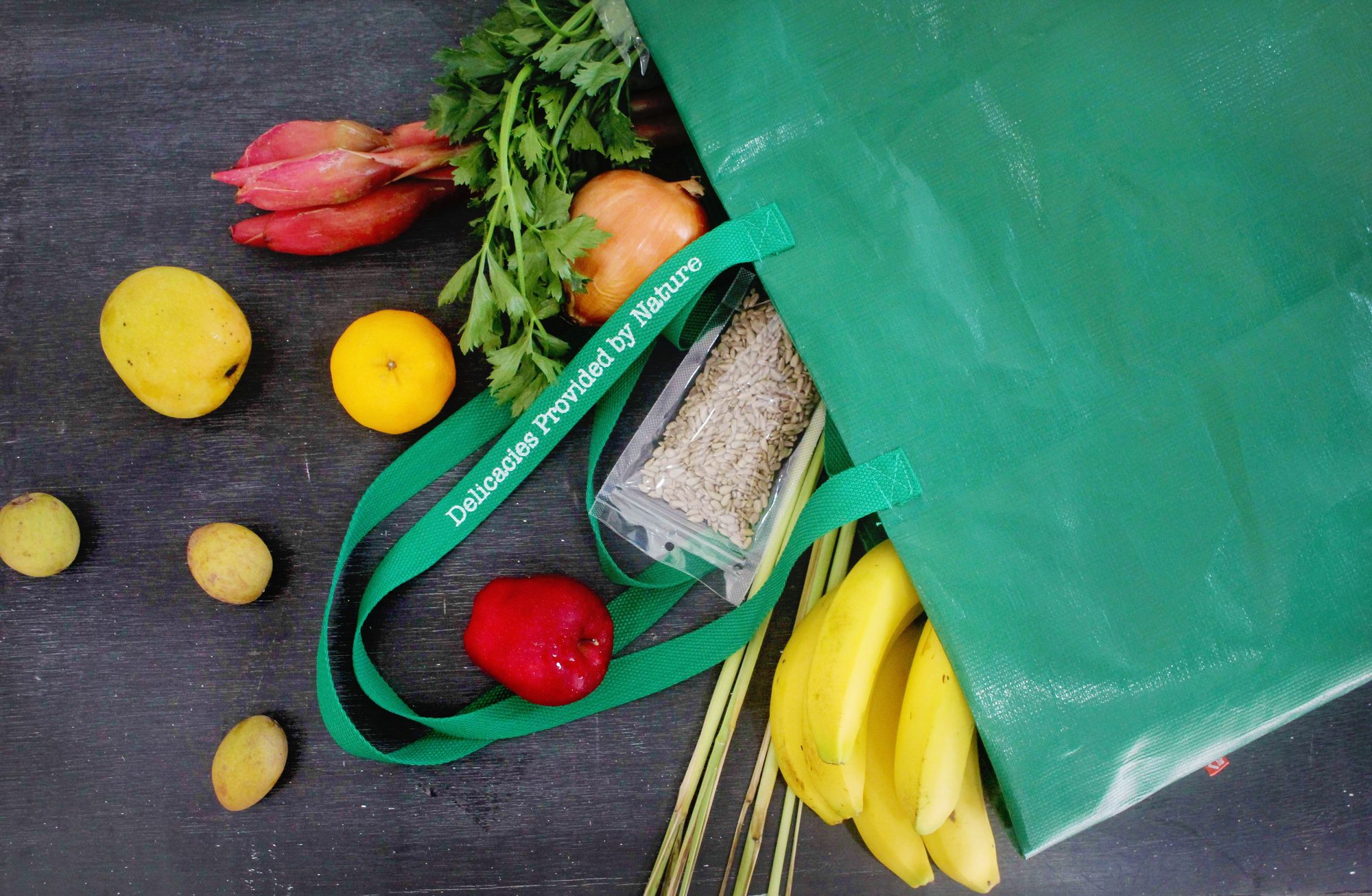 Top view of various fresh groceries in a green eco-friendly bag on a dark surface.