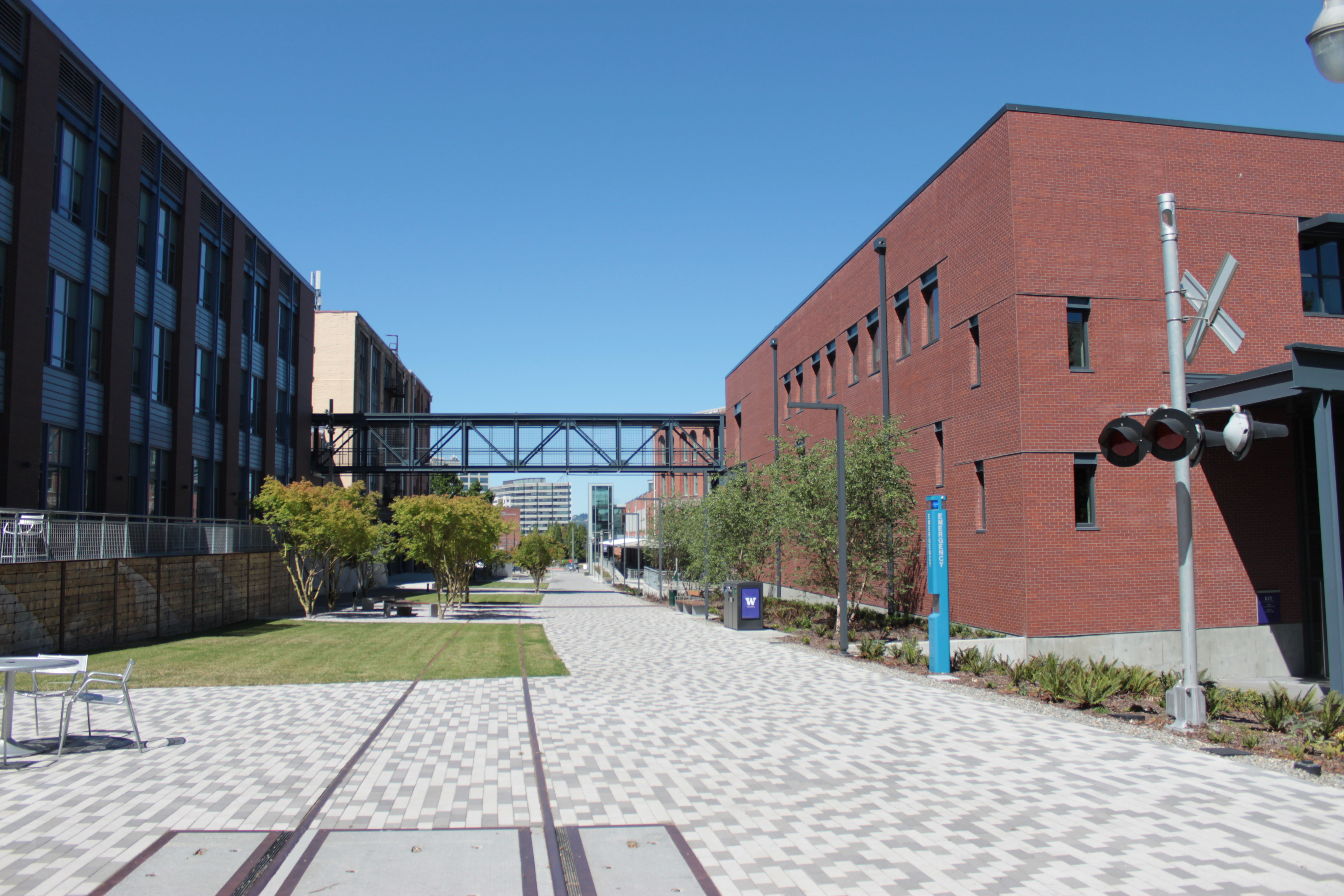 Brick-paved walkway with embedded rail tracks flanked by brick buildings and a steel pedestrian bridge on the UW Tacoma campus.
