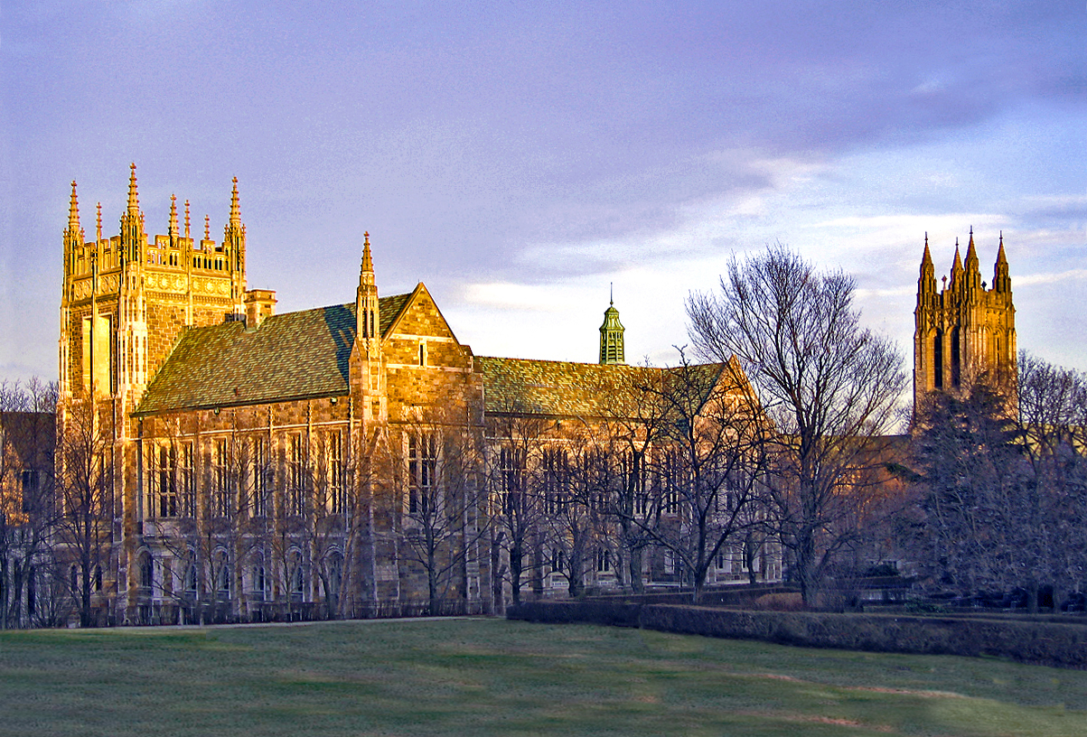 Gothic stone college buildings at sunset with bare trees and a green lawn in the foreground.