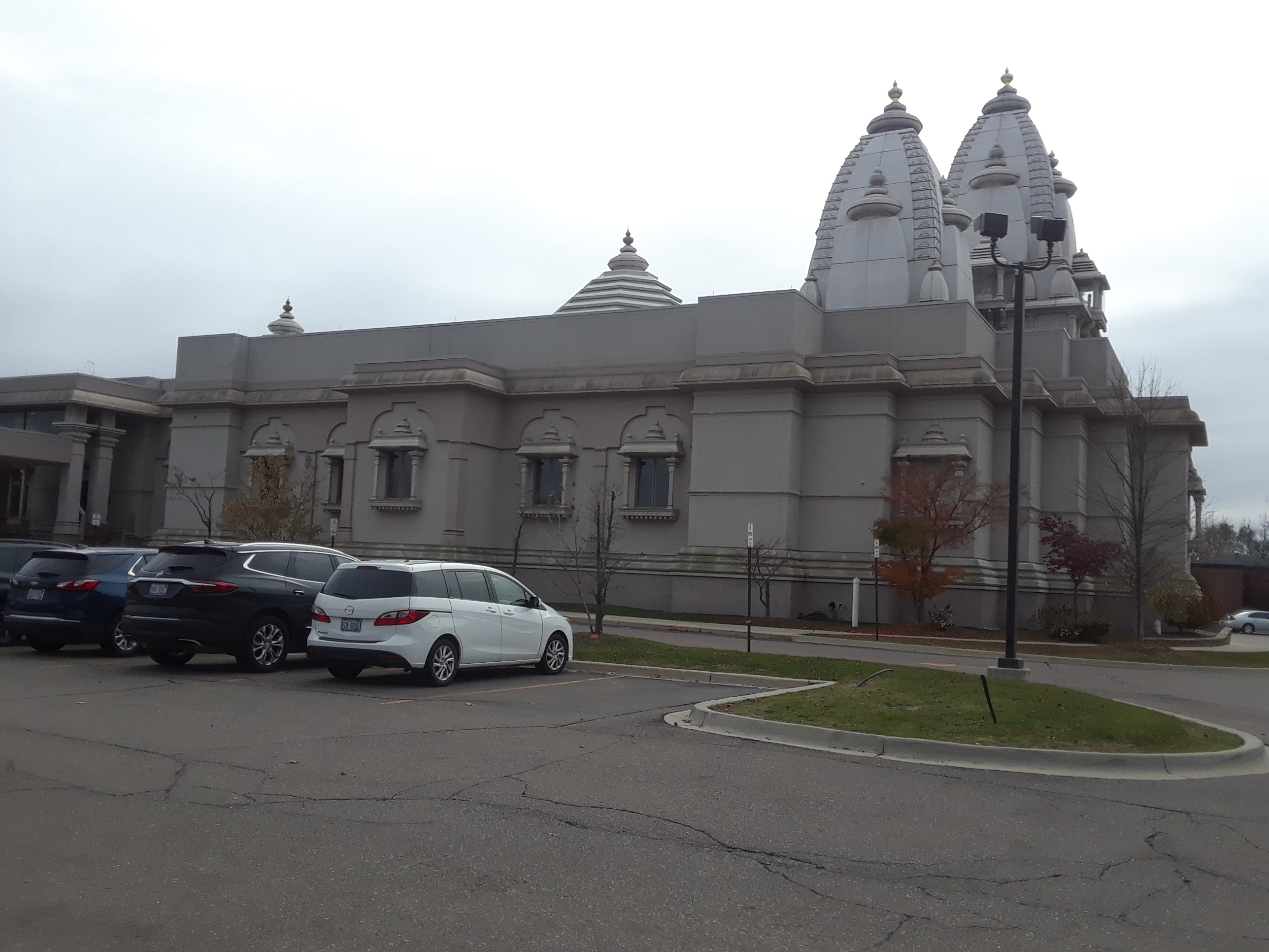 Hindu temple exterior with white ornate spires and a parking lot in front.