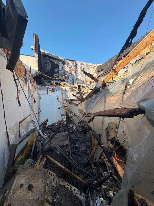Charred wooden beams and collapsed roof structure open to blue sky inside a fire-damaged building.