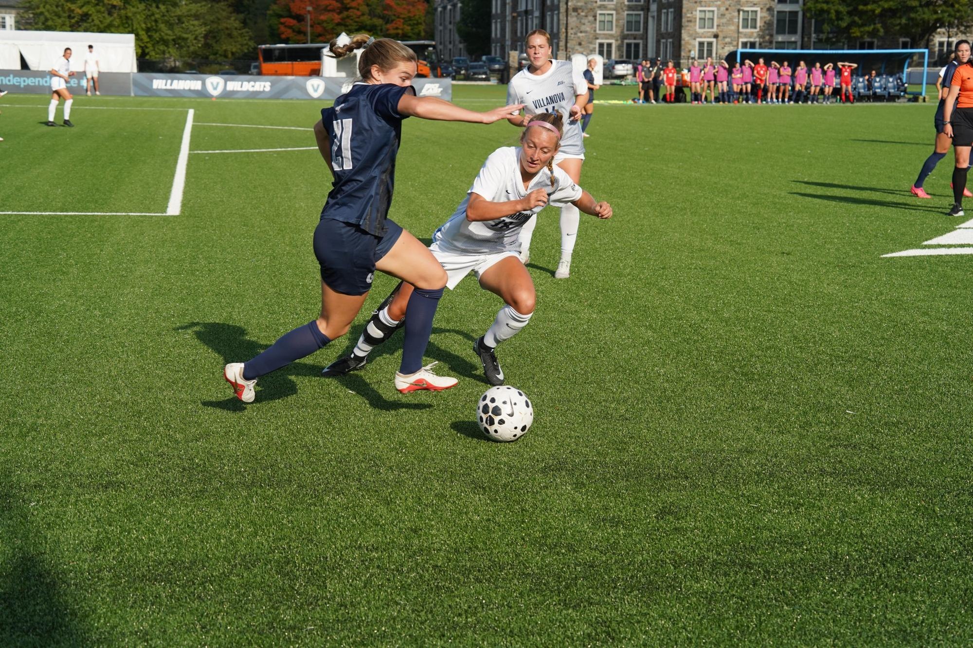 Two female soccer players in navy and white uniforms compete for the ball on a turf field.