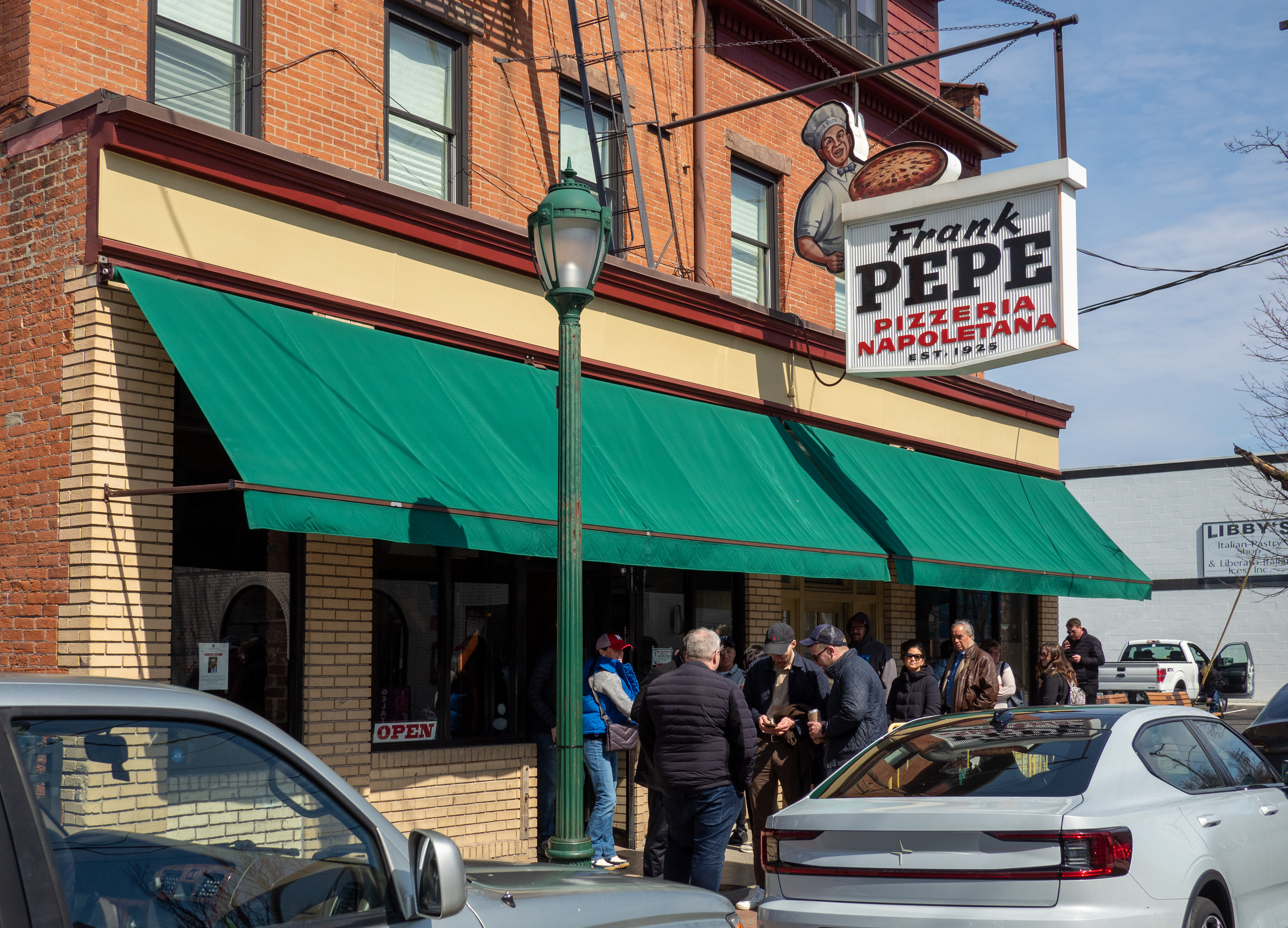 Brick restaurant exterior with green awnings, a Frank Pepe sign, and people waiting outside.
