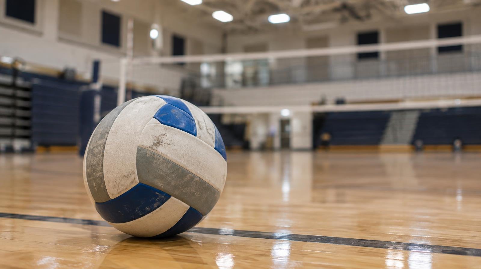 A volleyball sitting on a hardwood gym floor near the net with bleachers in the background.