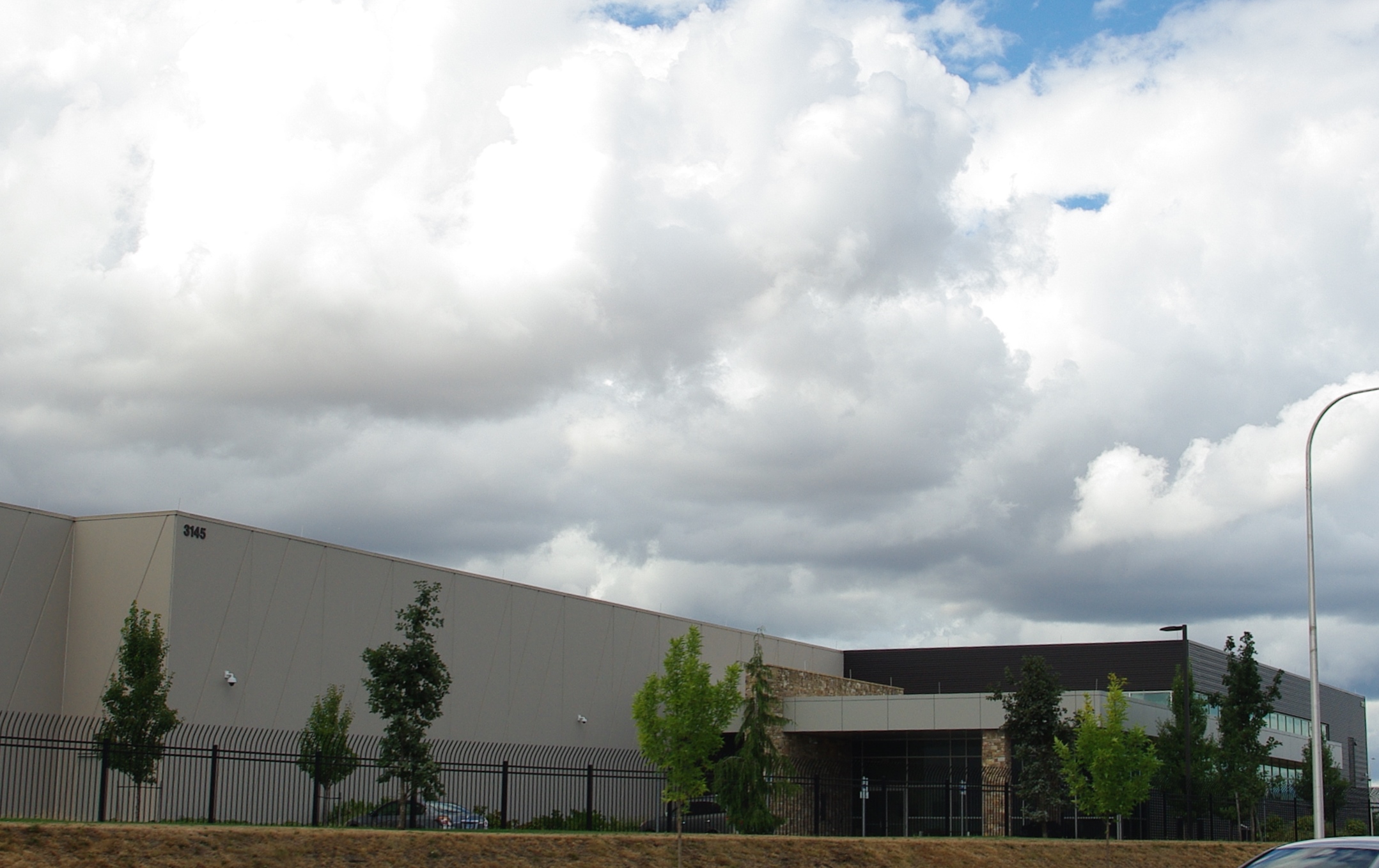 Modern data center building at a street corner with a fenced perimeter and partly cloudy sky.