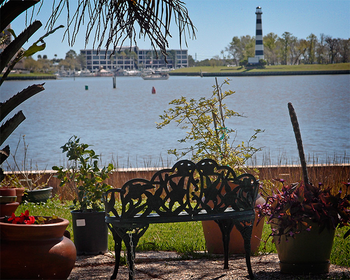 A bench overlooks calm water with a marina and striped lighthouse in the distance.