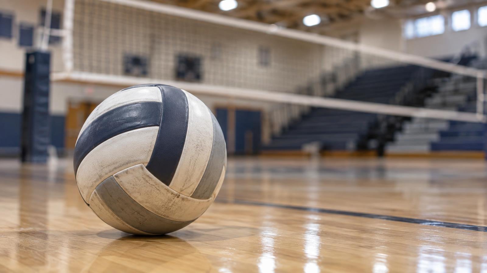 A volleyball resting on a hardwood gymnasium floor near a net with bleachers in the background.