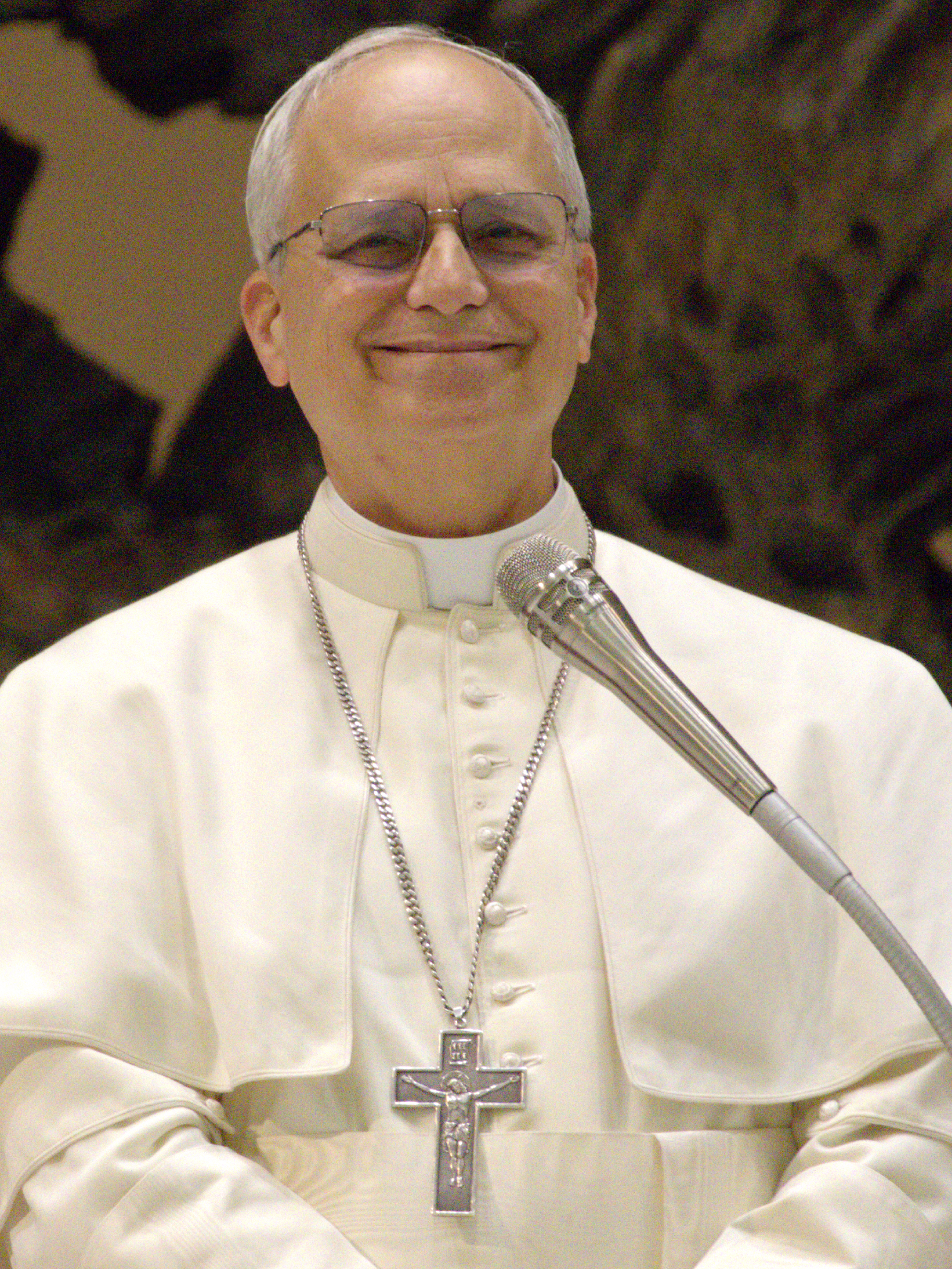 Smiling man in white papal vestments with a silver cross at a microphone.