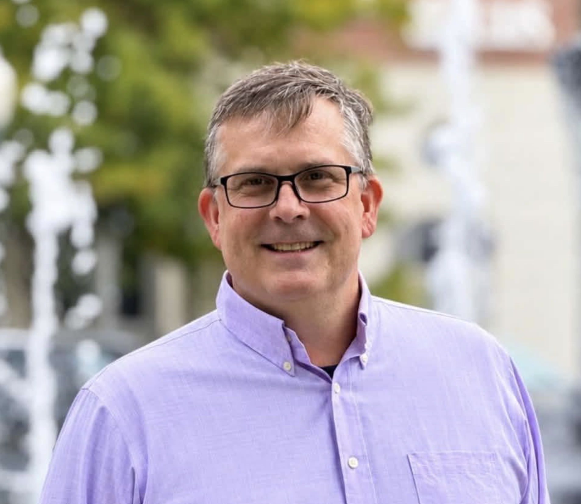 Dan Lemieux smiles outdoors, wearing black glasses and a light purple button-down shirt.