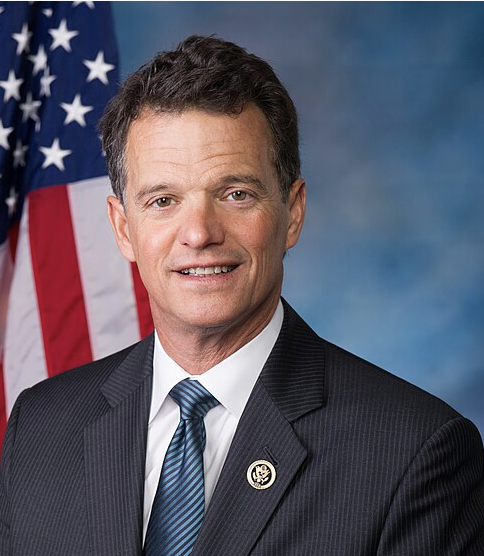 Former U.S. Rep. David Trott poses in a suit and tie in front of an American flag.