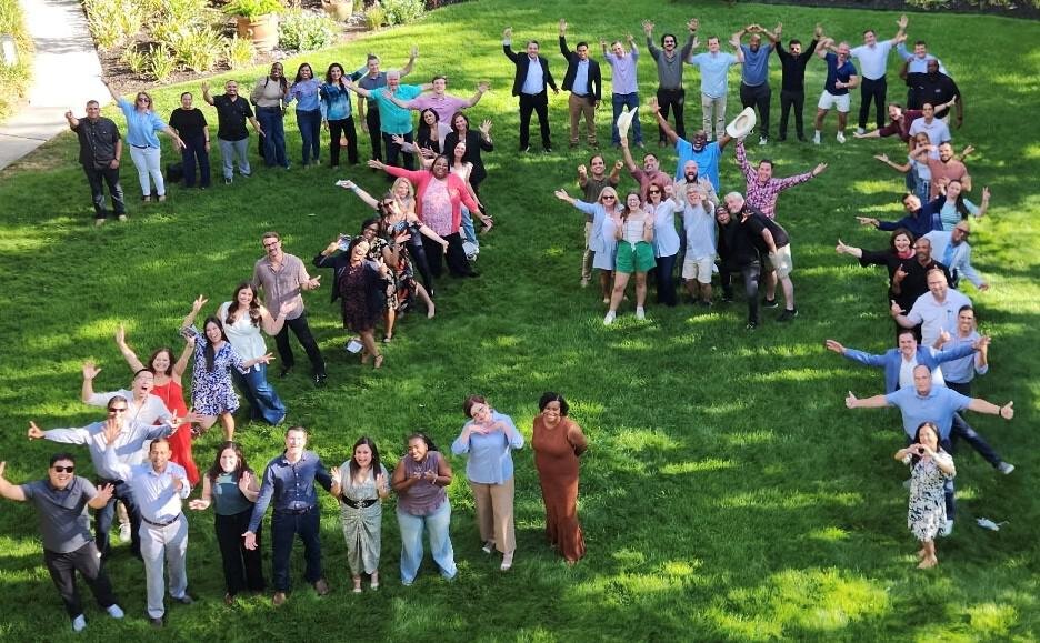 About 60 adults pose on a green lawn, many with arms raised, photographed from above.
