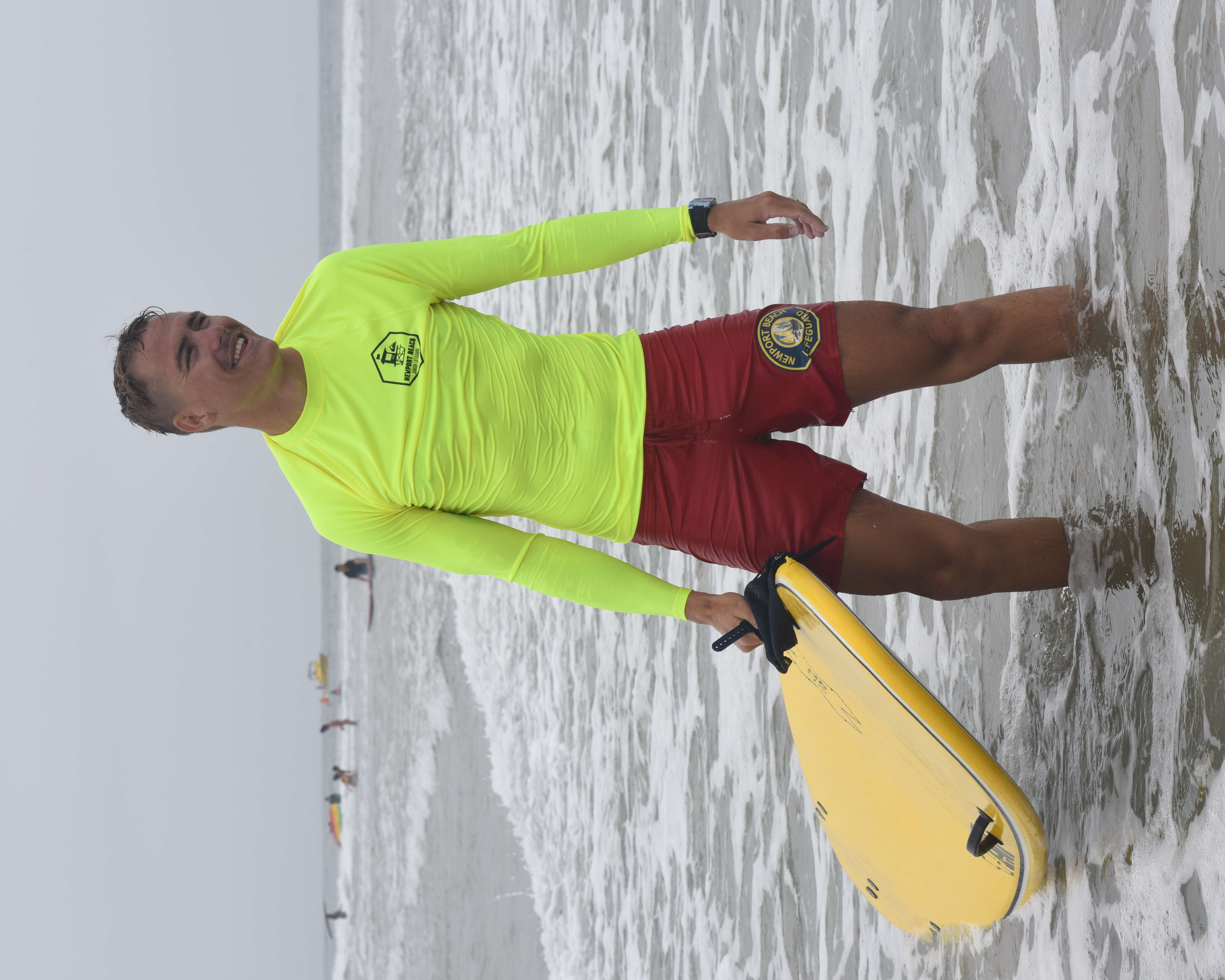 A lifeguard in a yellow rash guard standing in shallow ocean water holding a rescue buoy.