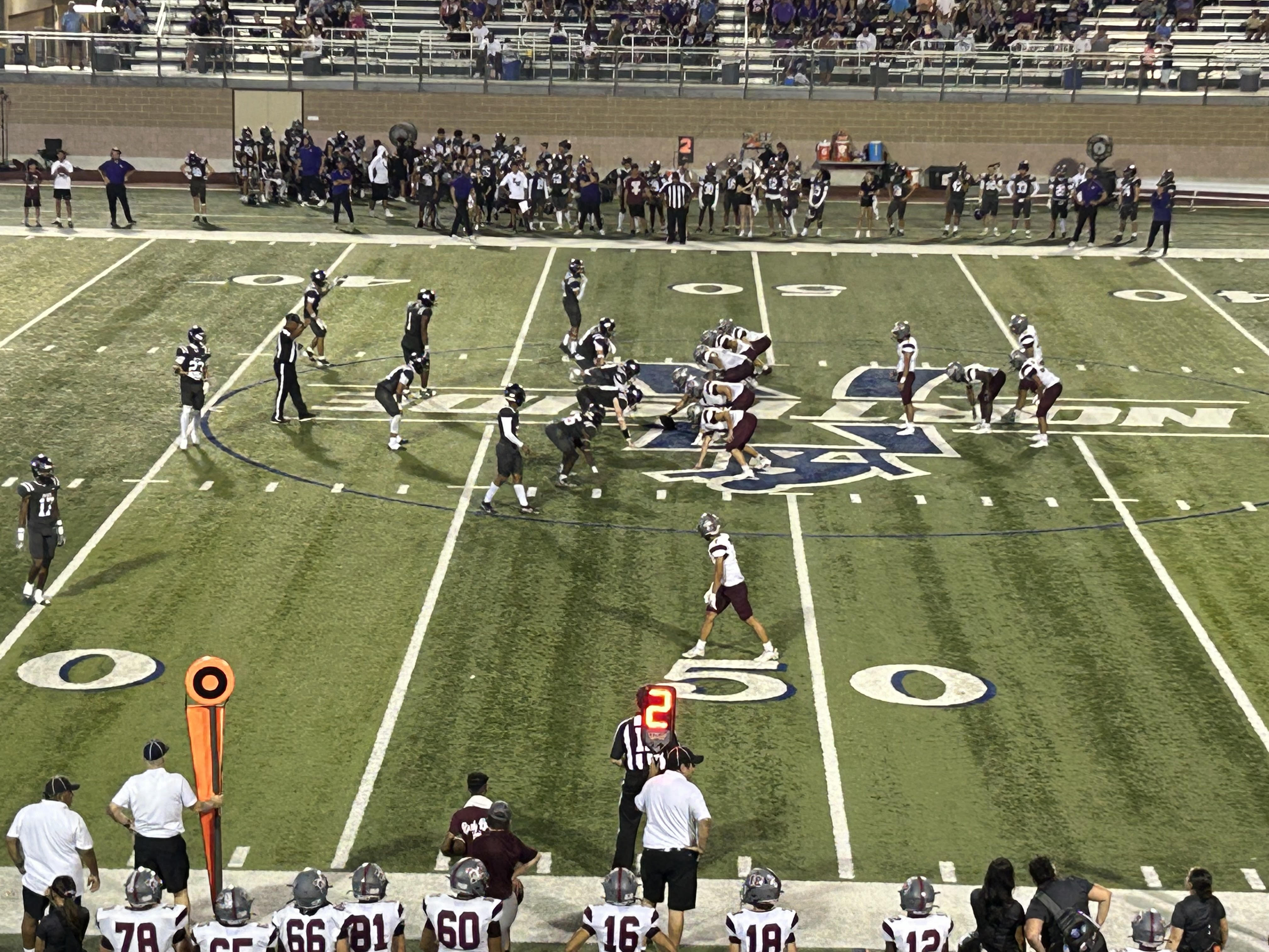 Overhead view of high school football players lined up at the line of scrimmage on a turf field at night.