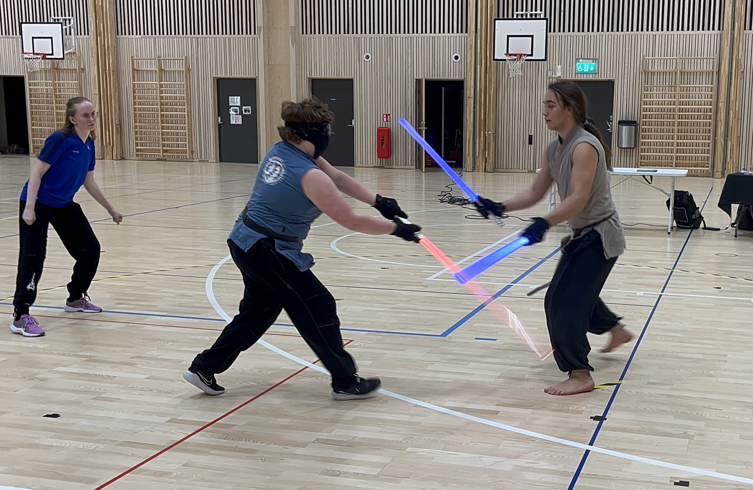 Two competitors duel with glowing blue and red lightsabers on a gym floor.