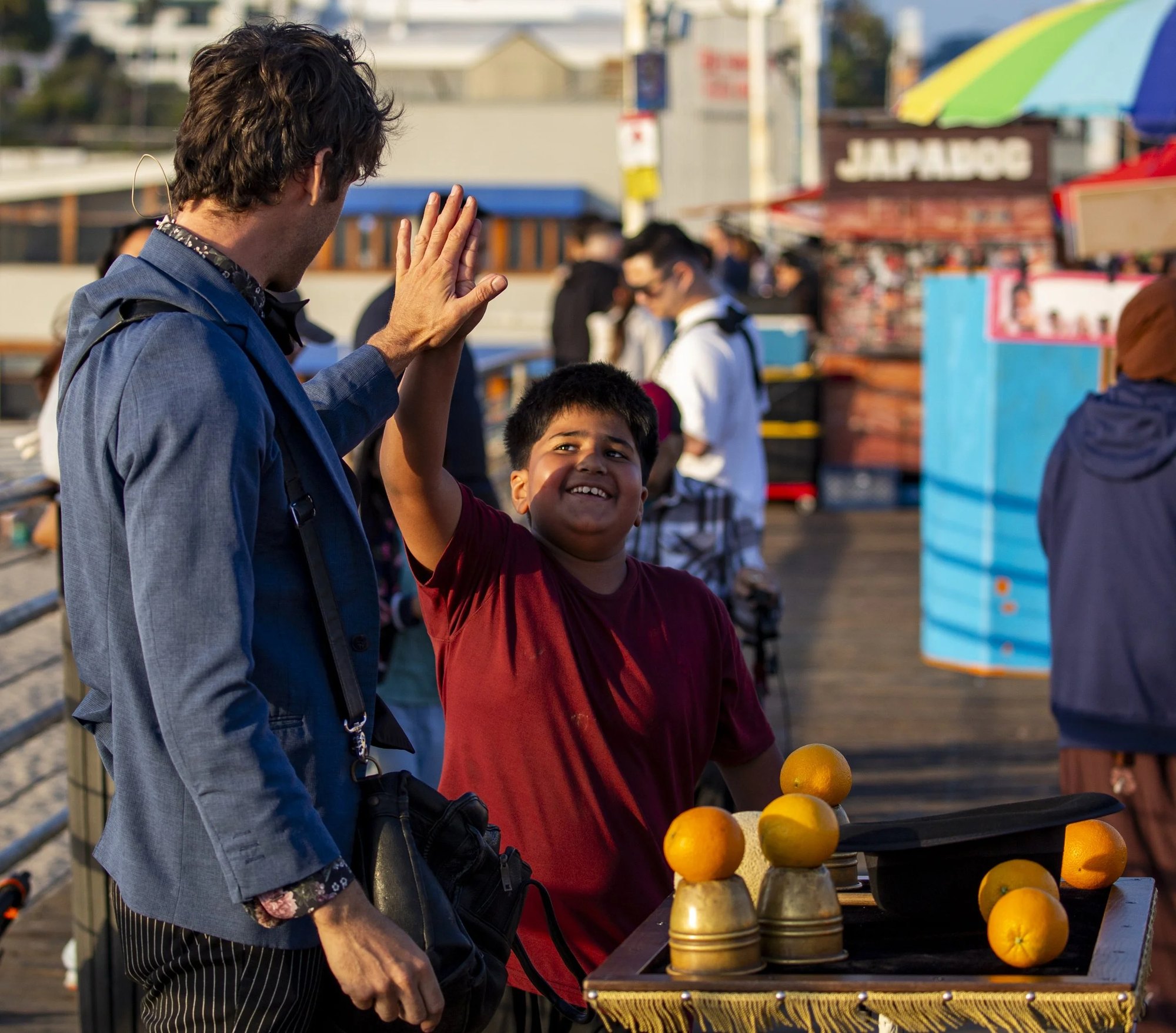 A man in a blue jacket high-fives a smiling boy on a boardwalk, with oranges and metal cups on a table in the foreground.