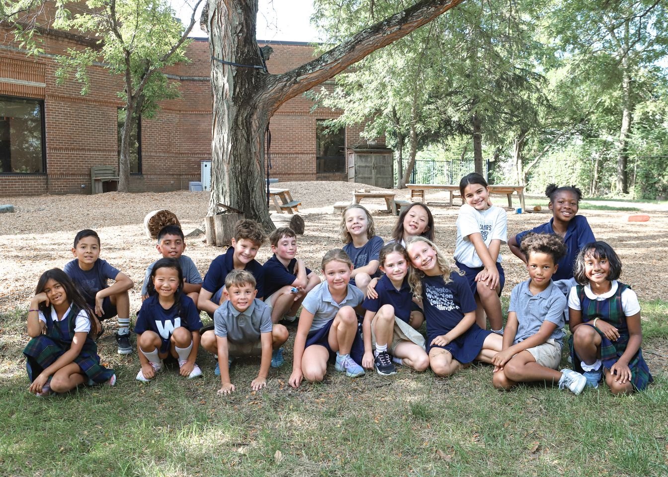 Group of elementary-aged children in navy uniforms posing outdoors in front of a brick building.
