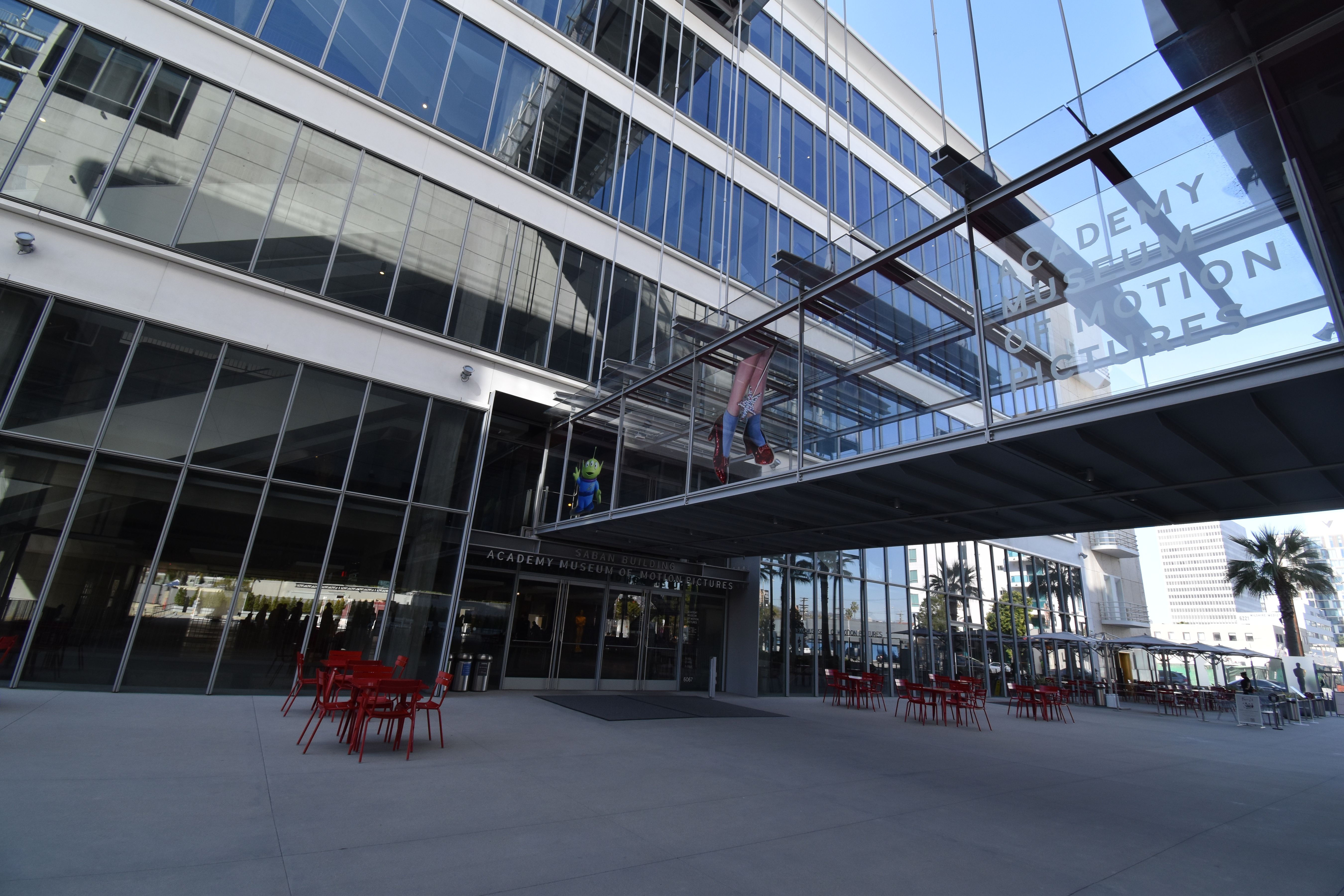 Glass-and-steel museum building exterior with an elevated walkway and red outdoor chairs.