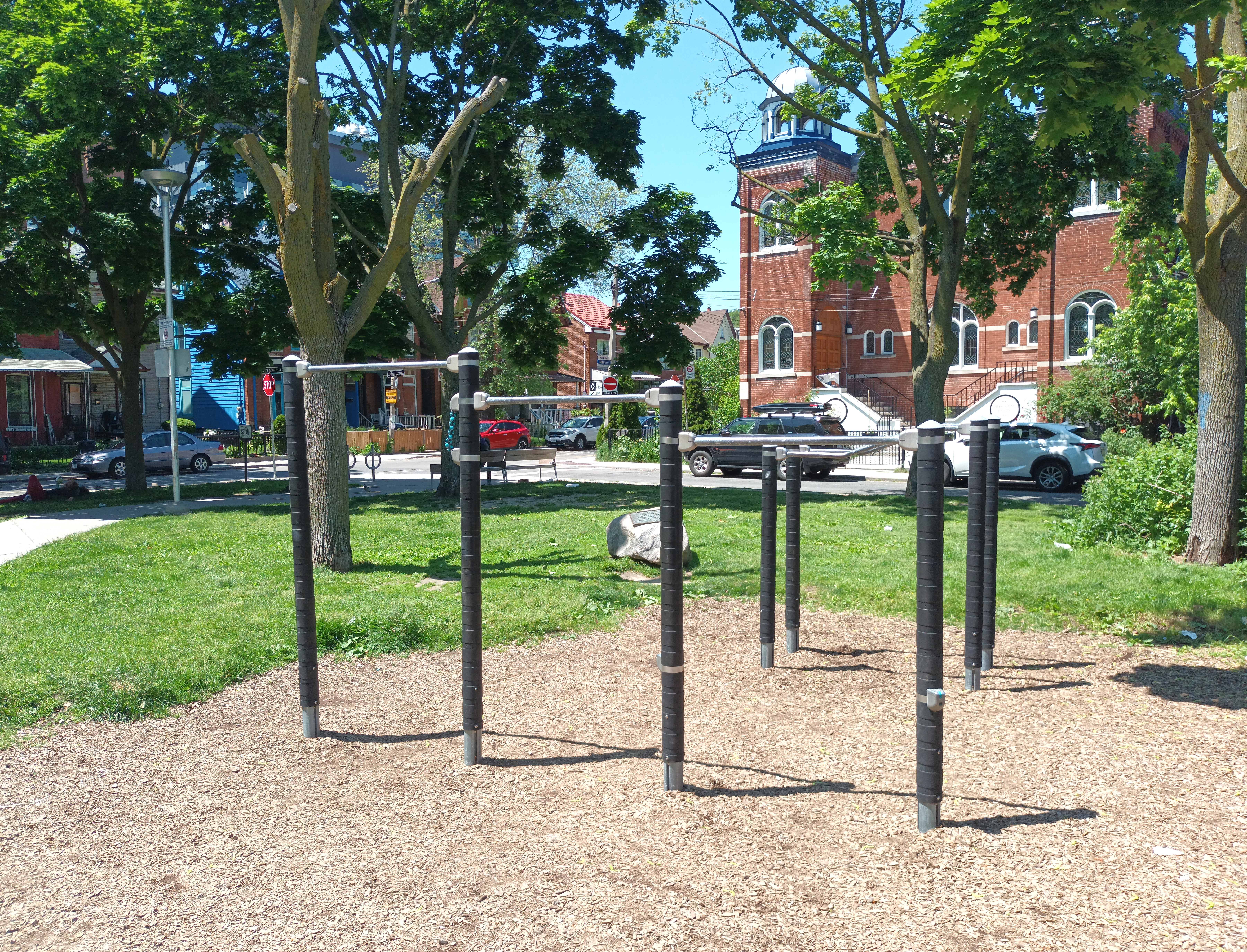 Metal exercise bars and fitness stations in a park with trees and wood-chip ground.