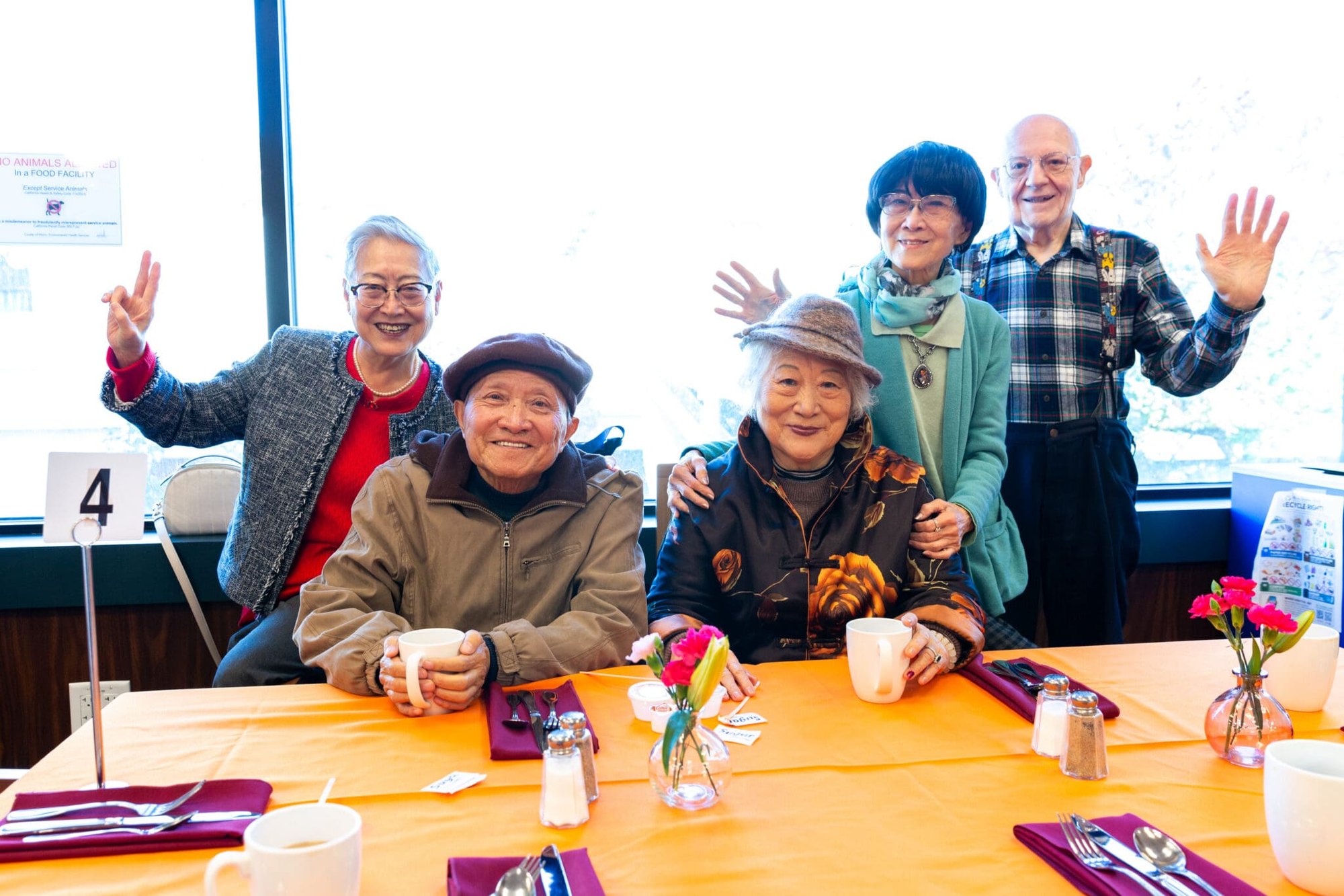 Six older adults smile and wave at a table decorated with orange cloth and small flowers.