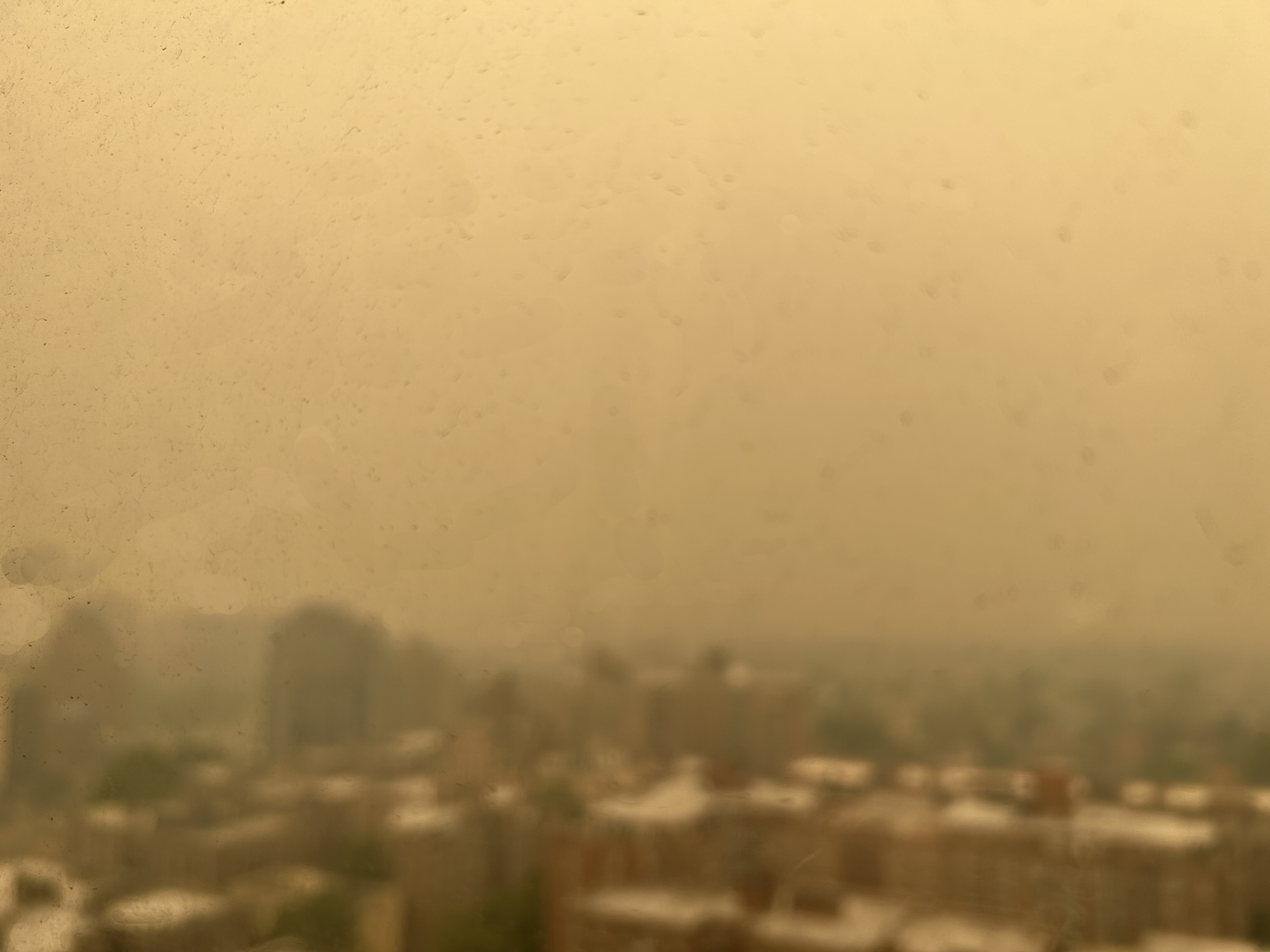 City skyline barely visible through thick yellowish-brown haze, seen through a window.