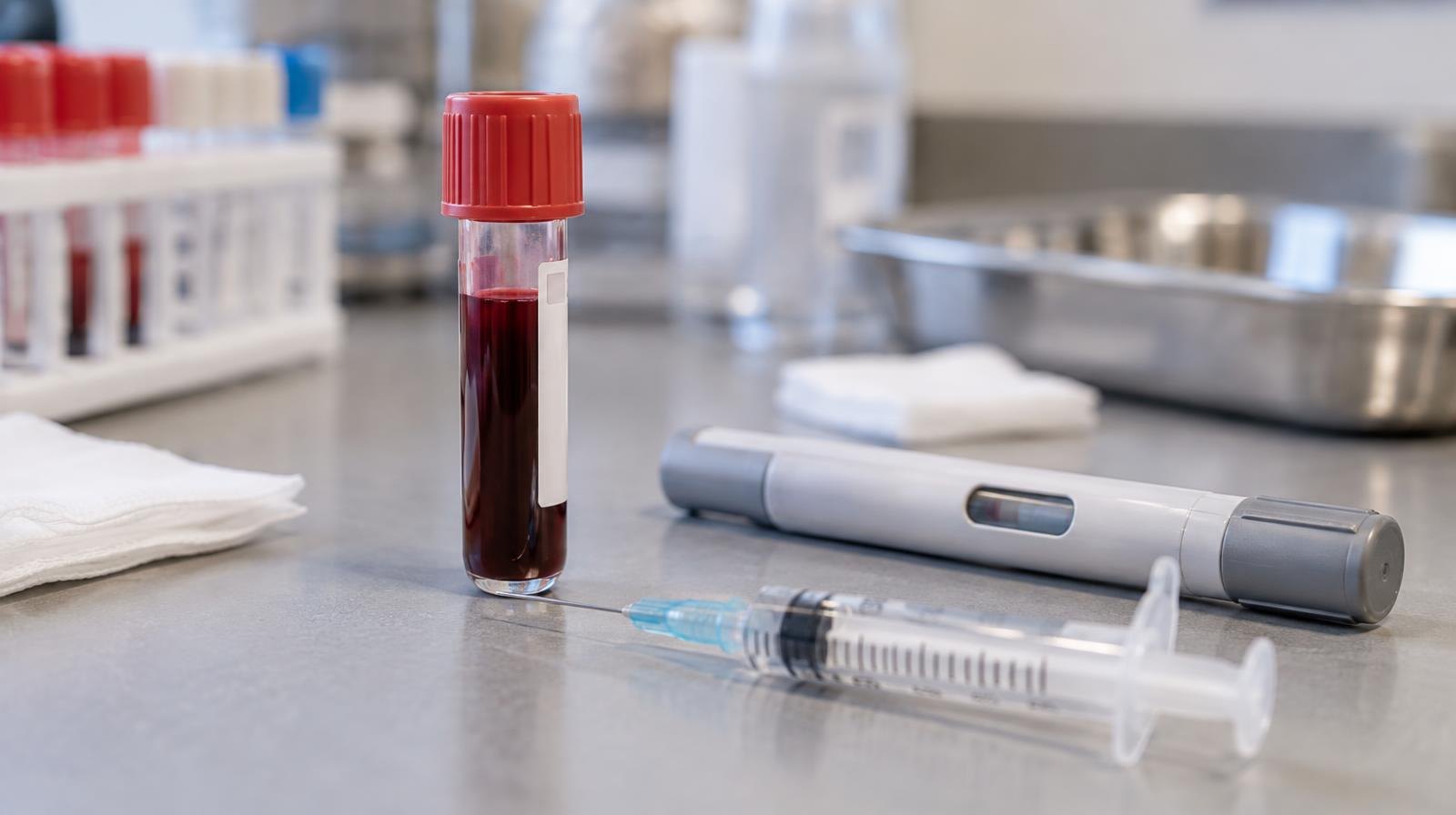 A glass blood vial, syringe, and medical injection pen on a lab surface.