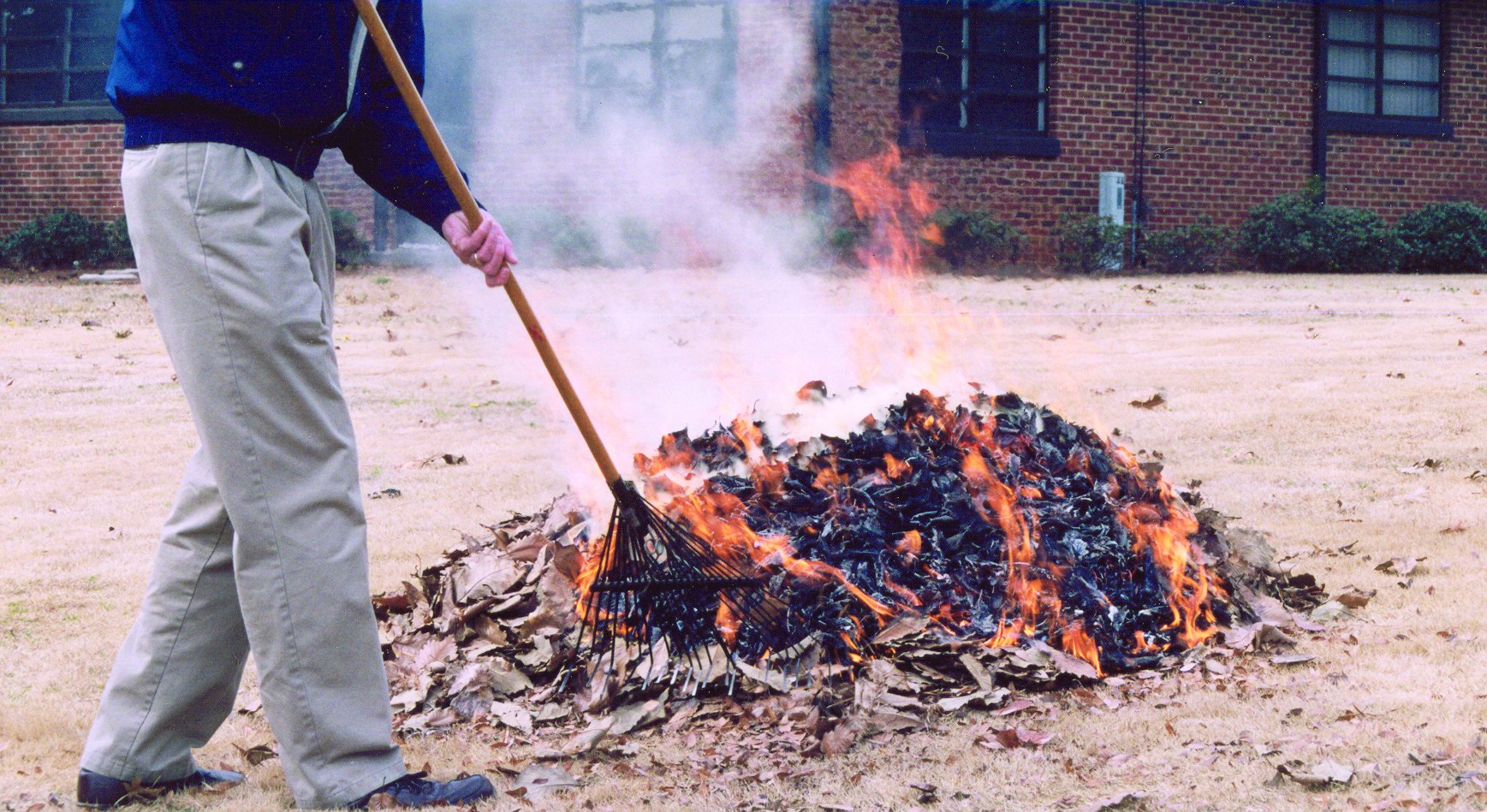 Person in a blue jacket raking a large burning pile of leaves and yard debris outdoors.
