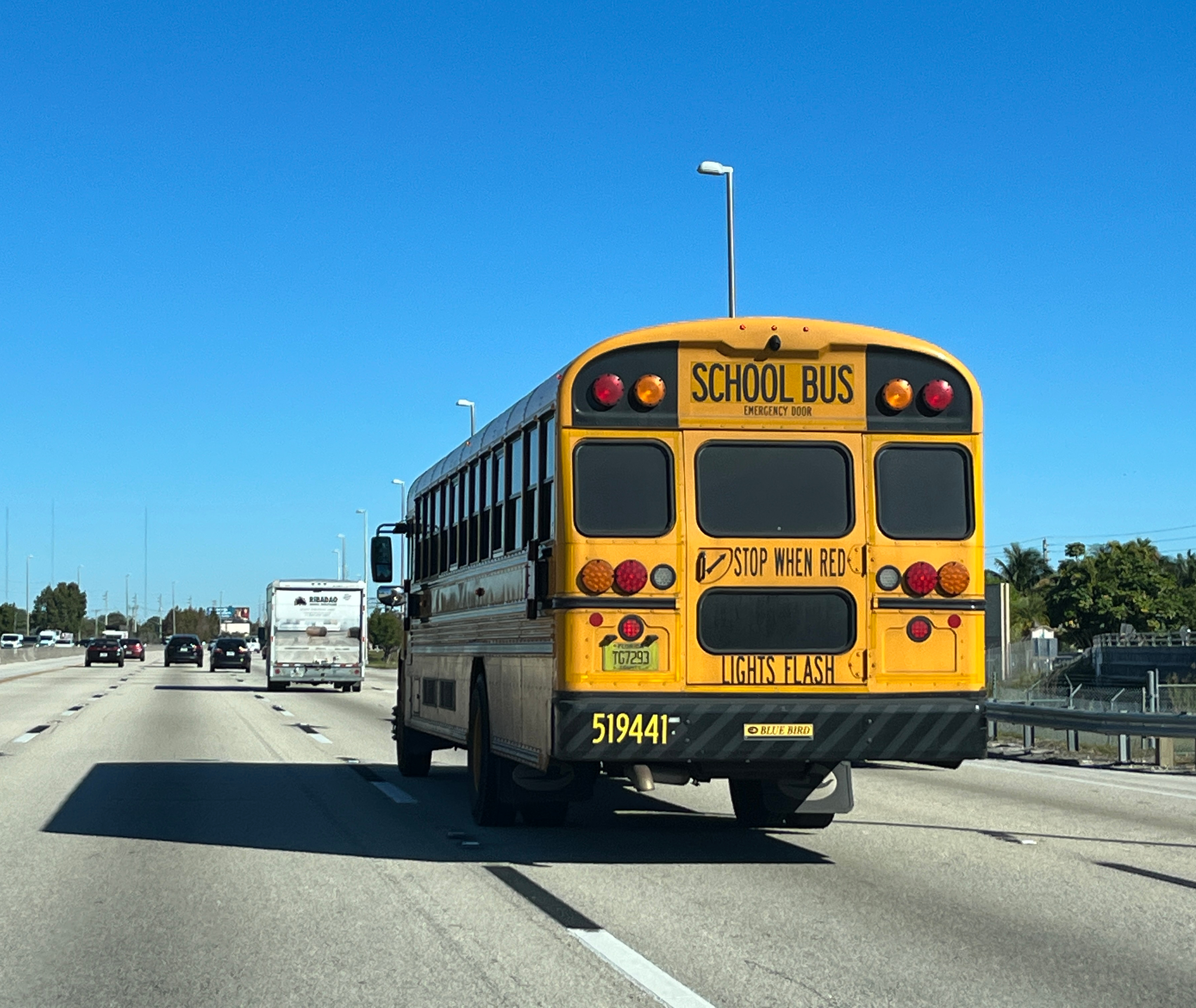 Rear of a yellow school bus traveling on a multi-lane highway under a clear blue sky.