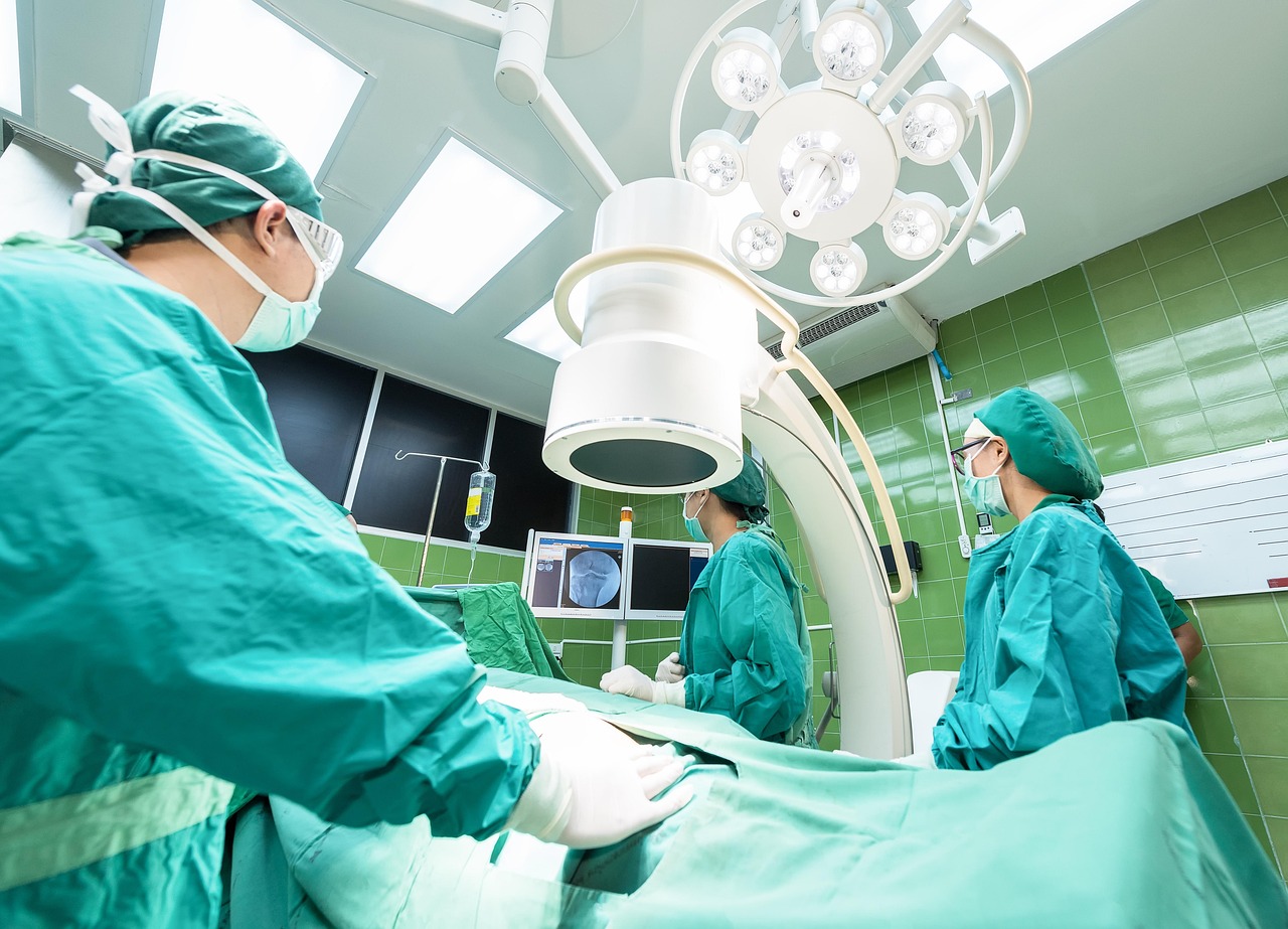 Three medical staff members in green surgical attire stand around a draped operating table beneath surgical lights and imaging equipment.