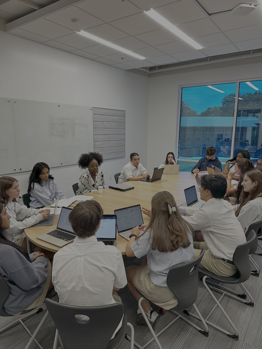 Students seated around an oval conference table with laptops open in a modern meeting room.
