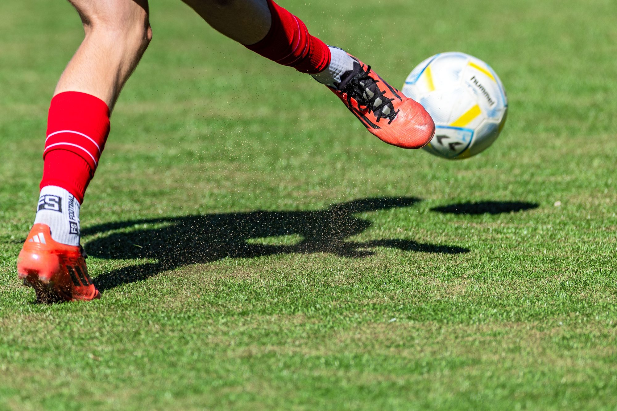 Soccer player in red kicks a ball mid-air on a sunny day outdoor field.