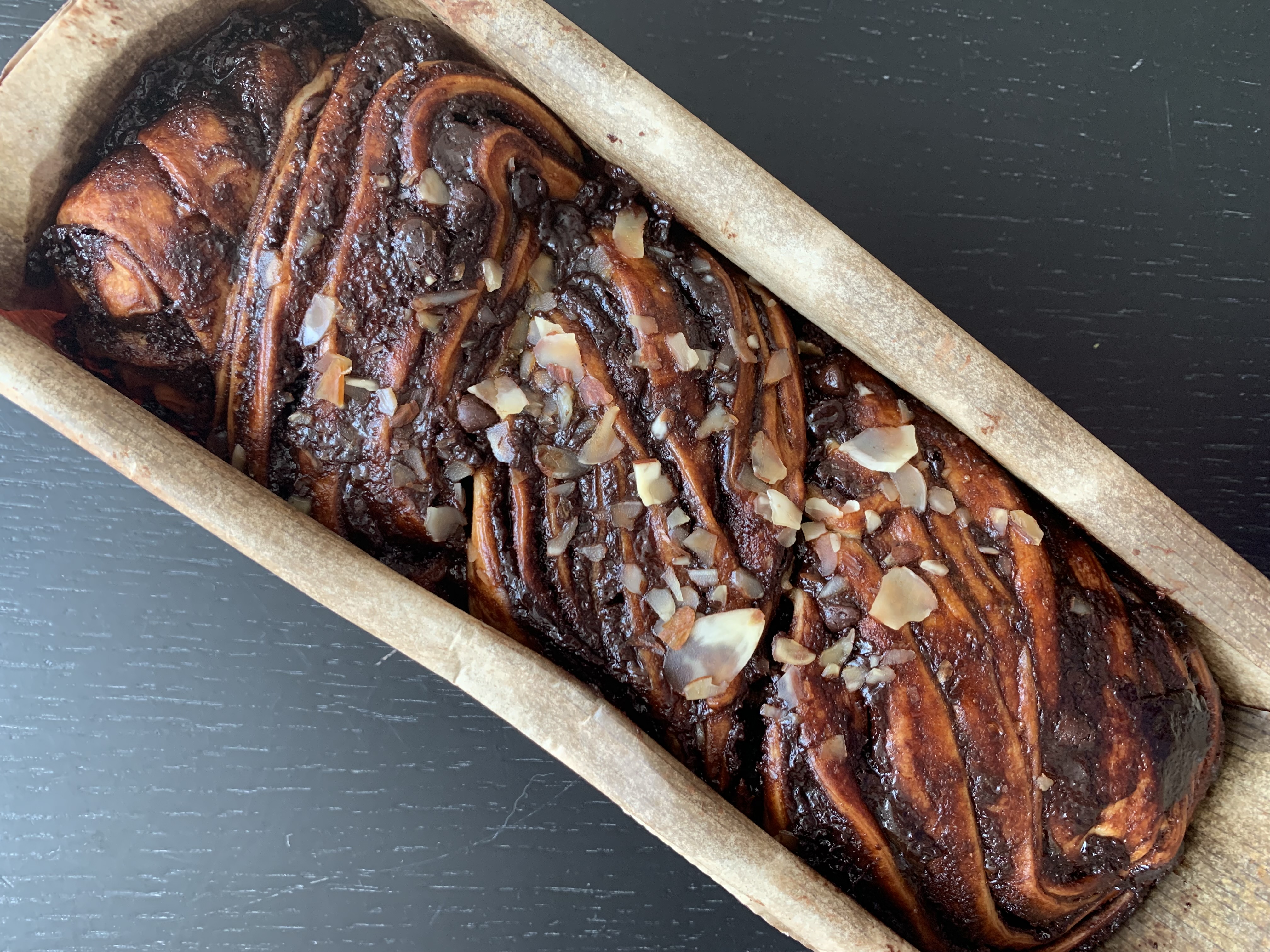 A chocolate babka loaf with twisted dough and almond topping in a parchment-lined mold.