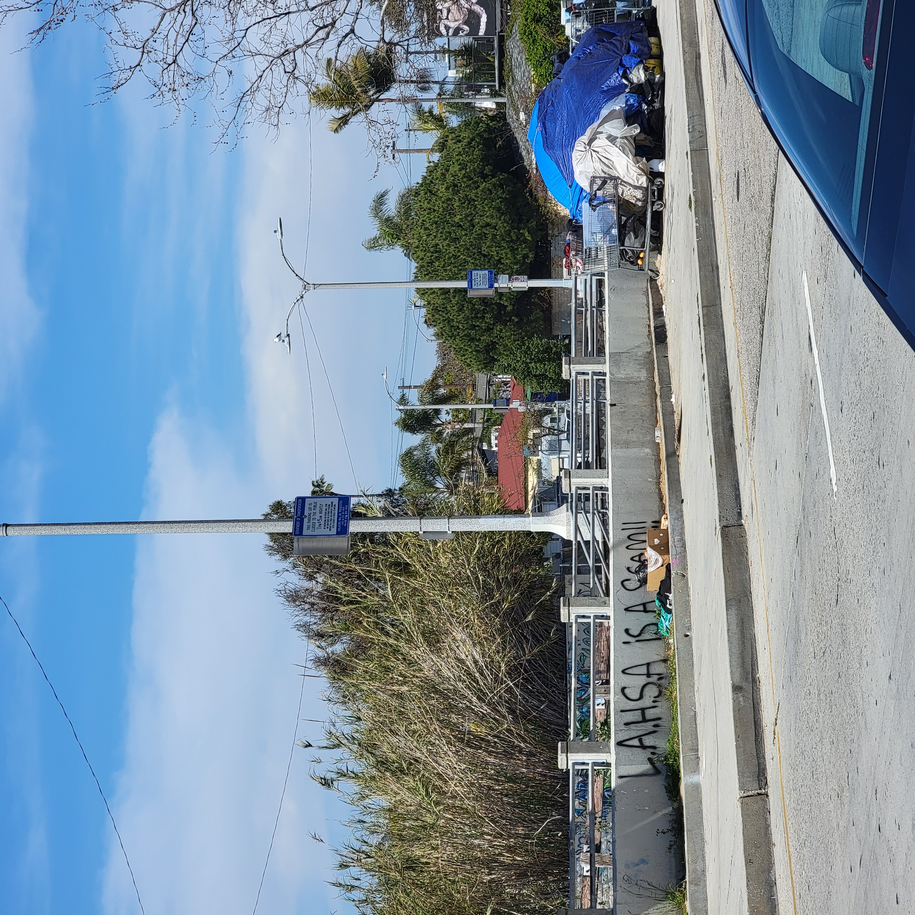 Graffiti on a concrete barrier reads 'LAHSA IS A SCAM' with a blue-tarp encampment behind it.