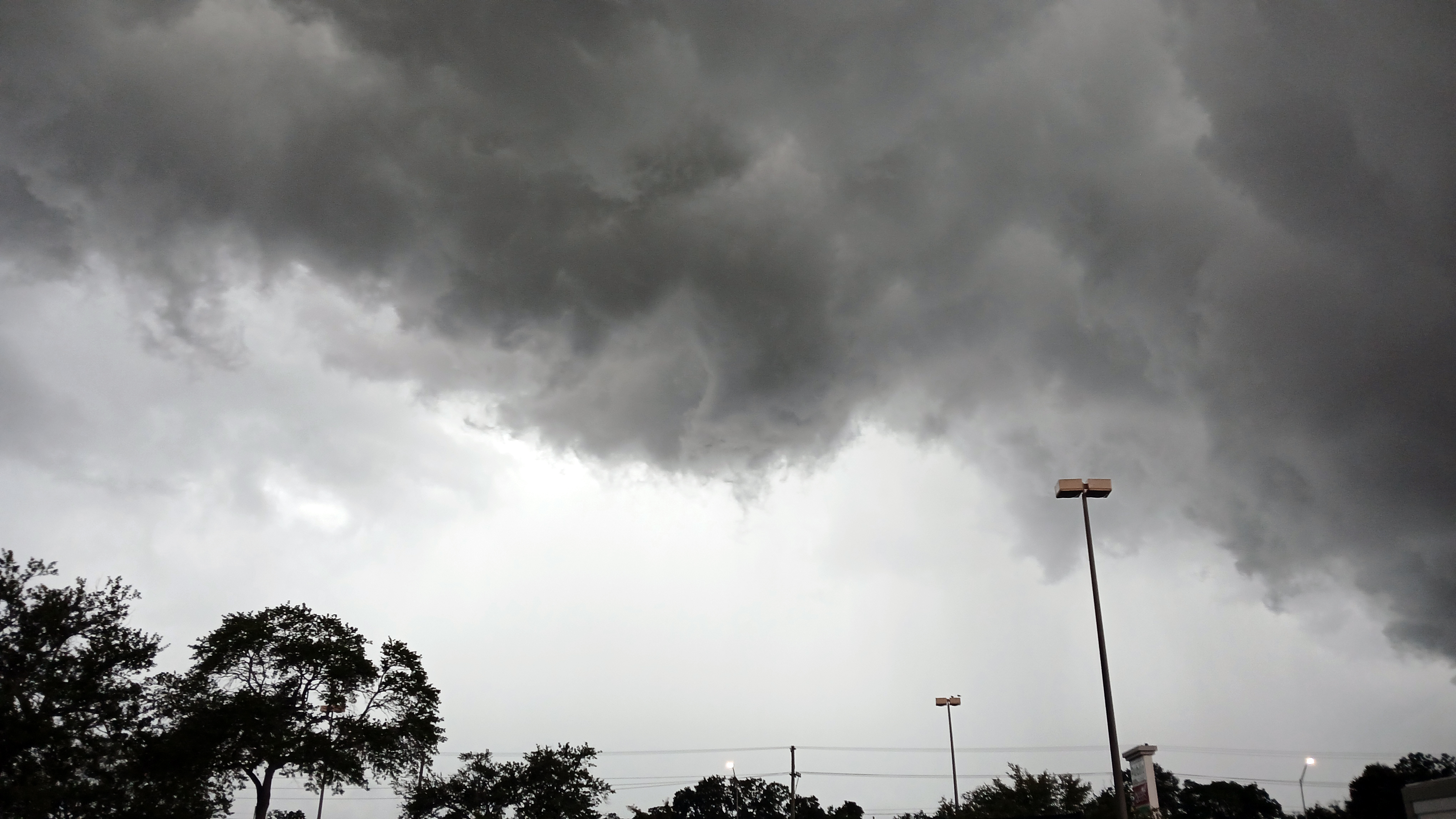 Dark grey storm clouds fill the sky above a parking lot with street lights and trees.