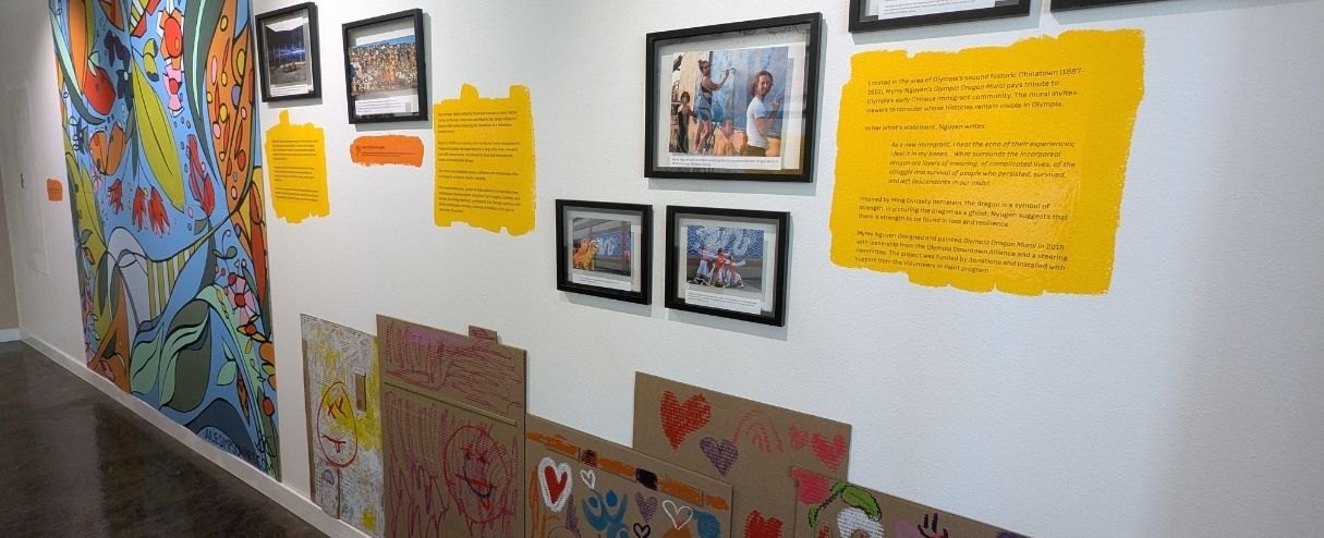 Museum gallery wall with framed photos, yellow text panels, a large mural, and children's drawings on cardboard.
