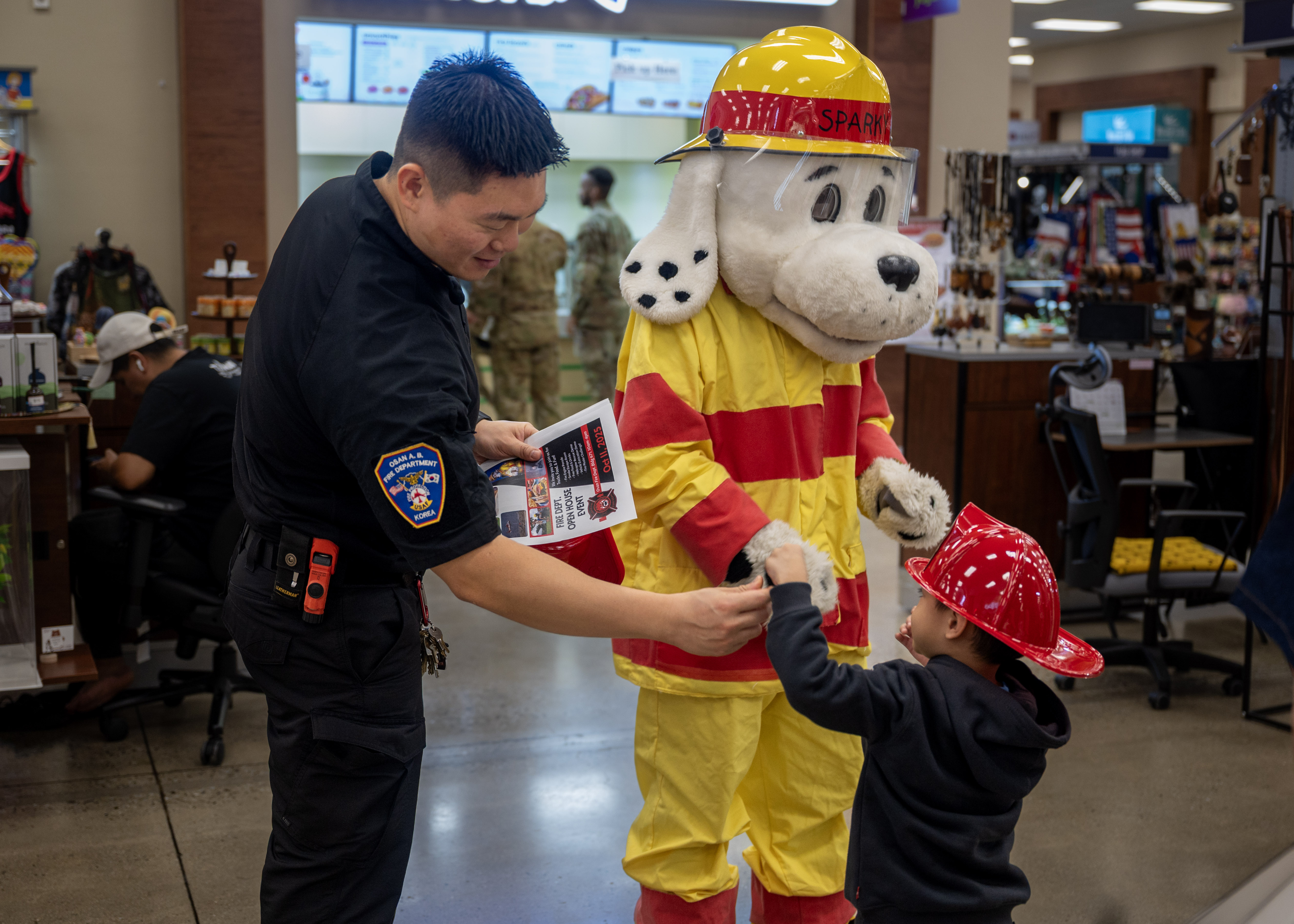 A firefighter and a mascot in a fire dog costume give a badge to a child in a toy helmet.