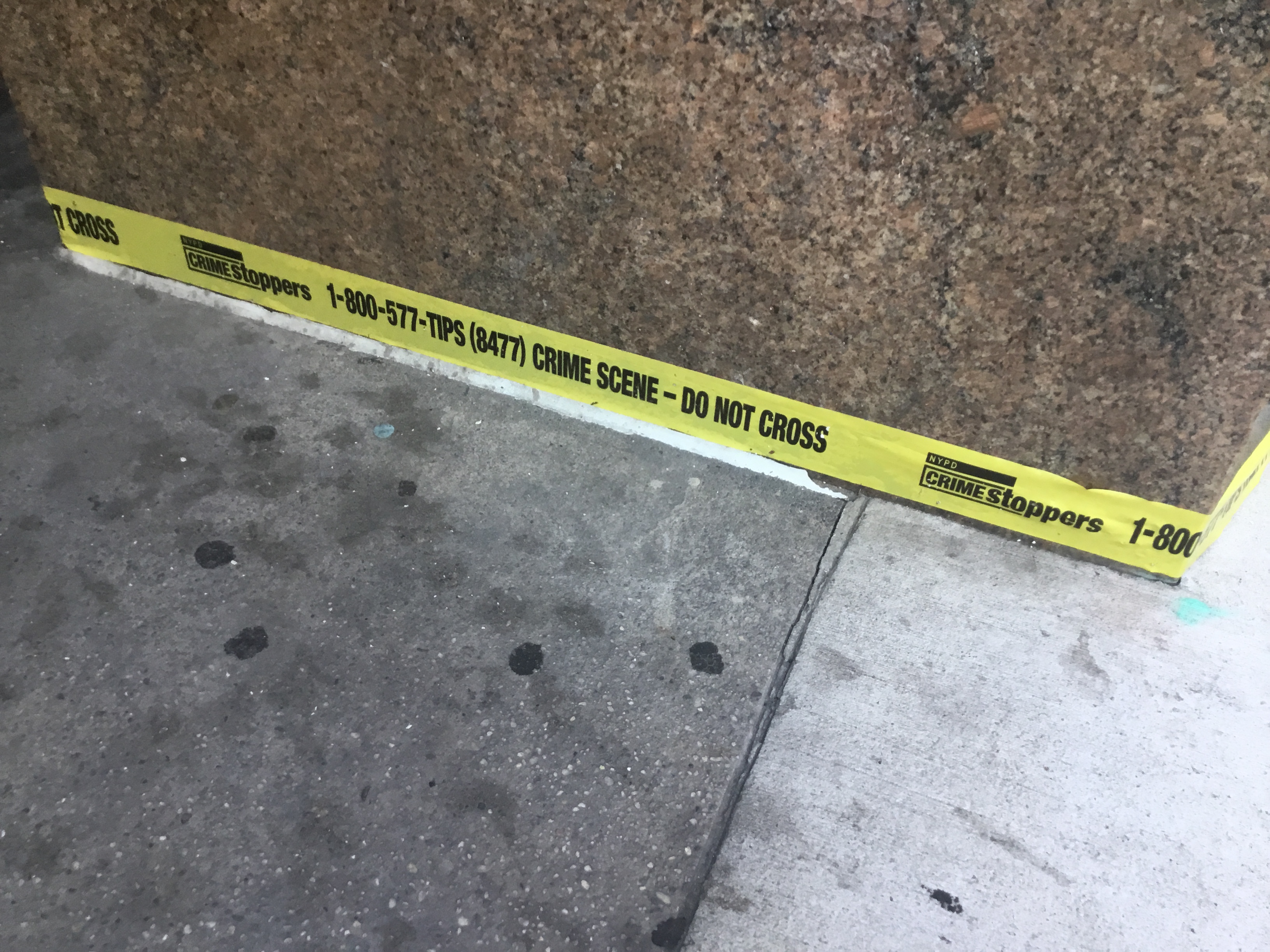 Yellow crime scene tape reading 'Do Not Cross' stretched along a stone wall on a sidewalk.