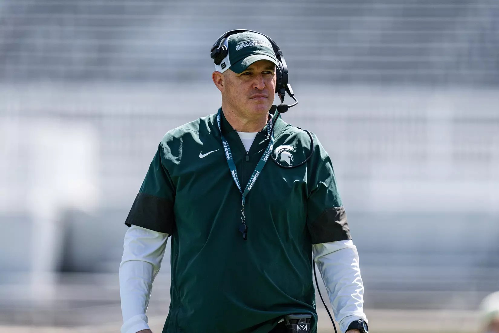 A man in a green Michigan State coaching shirt and headset walks on a football field.
