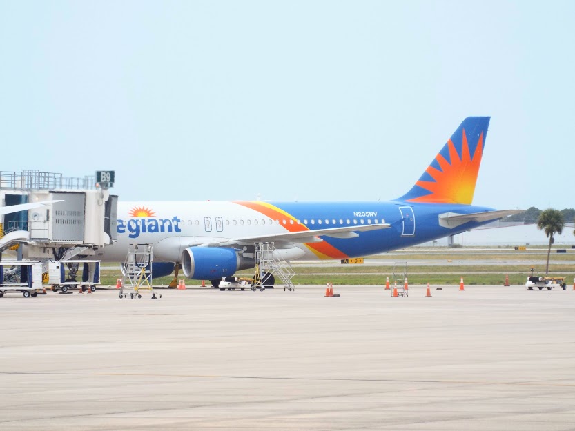 Allegiant Air blue-and-orange jet parked at an airport gate with jet bridge attached.