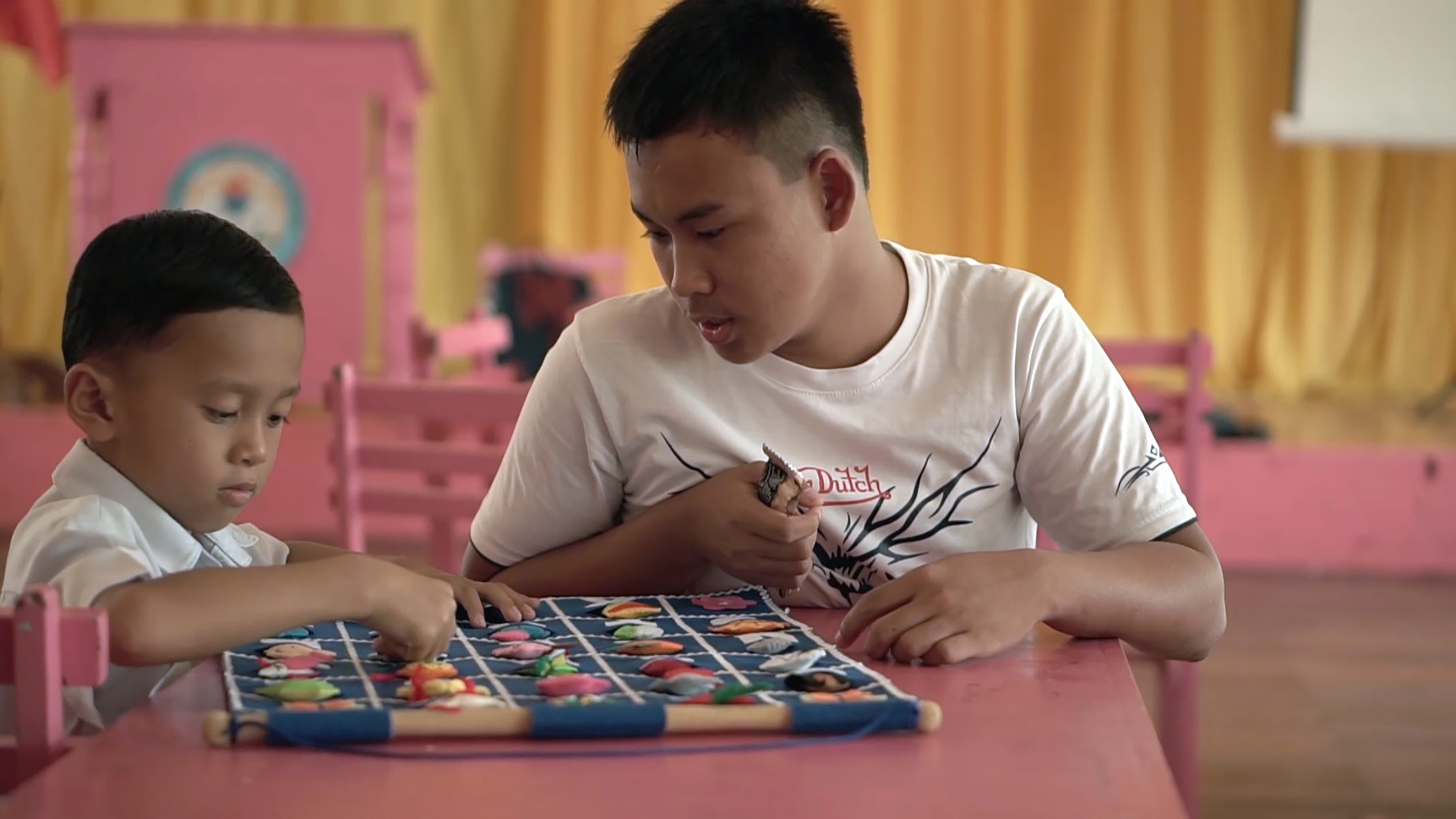 Two young boys sit at a classroom table, one helping the other with a learning activity.