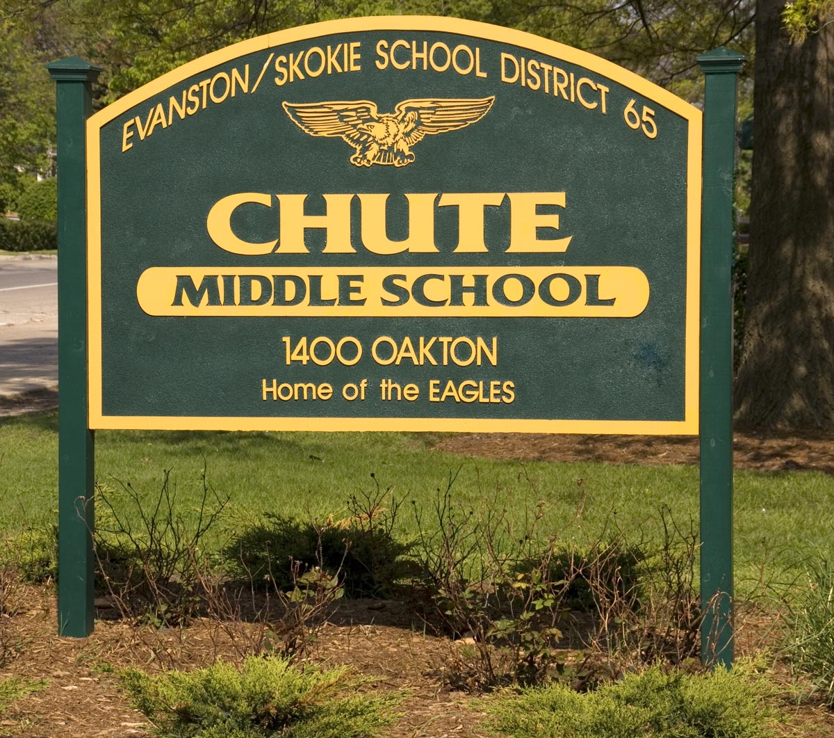 Green and gold sign for Chute Middle School displaying District 65 affiliation, mounted on posts near trees.