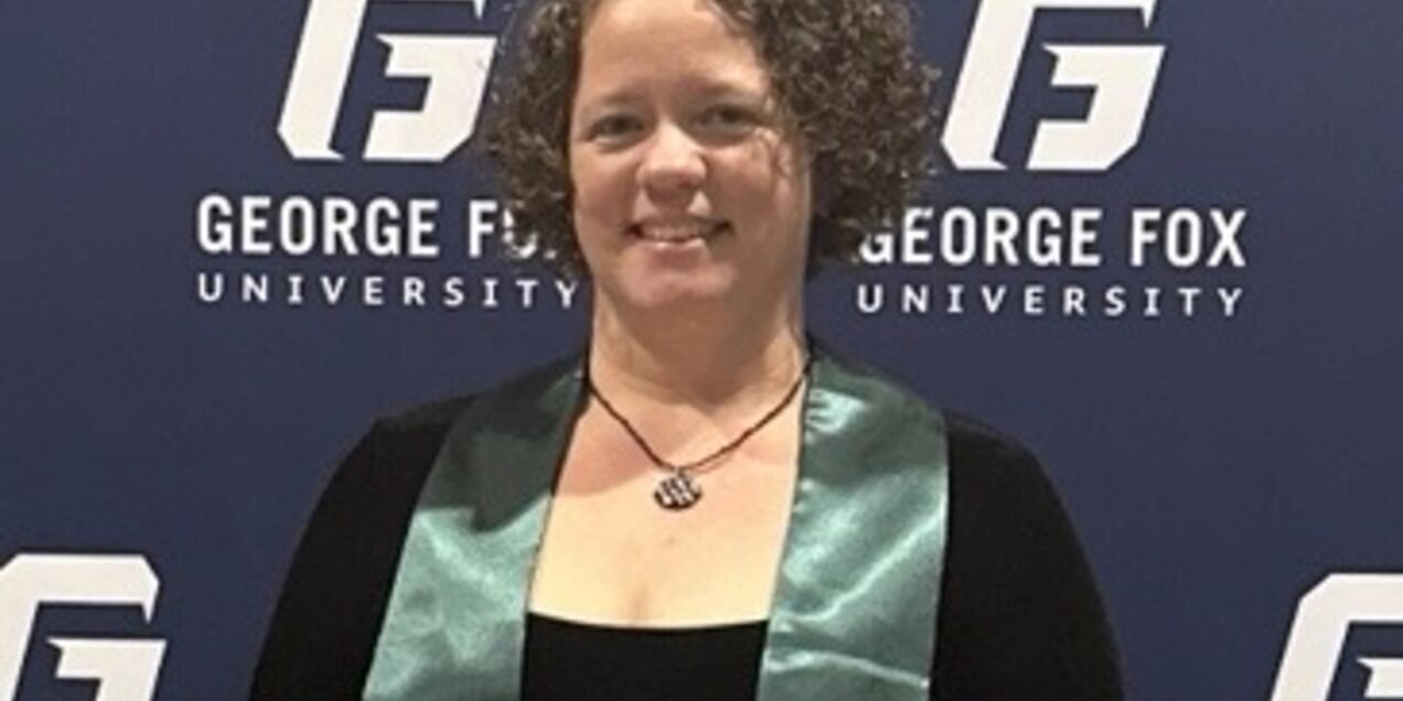 Woman in graduation sash smiling in front of a university branded backdrop.